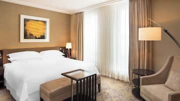 Quarto de hotel com uma cama grande, mesa de cabeceira com abajur, uma cadeira e um quadro na parede.