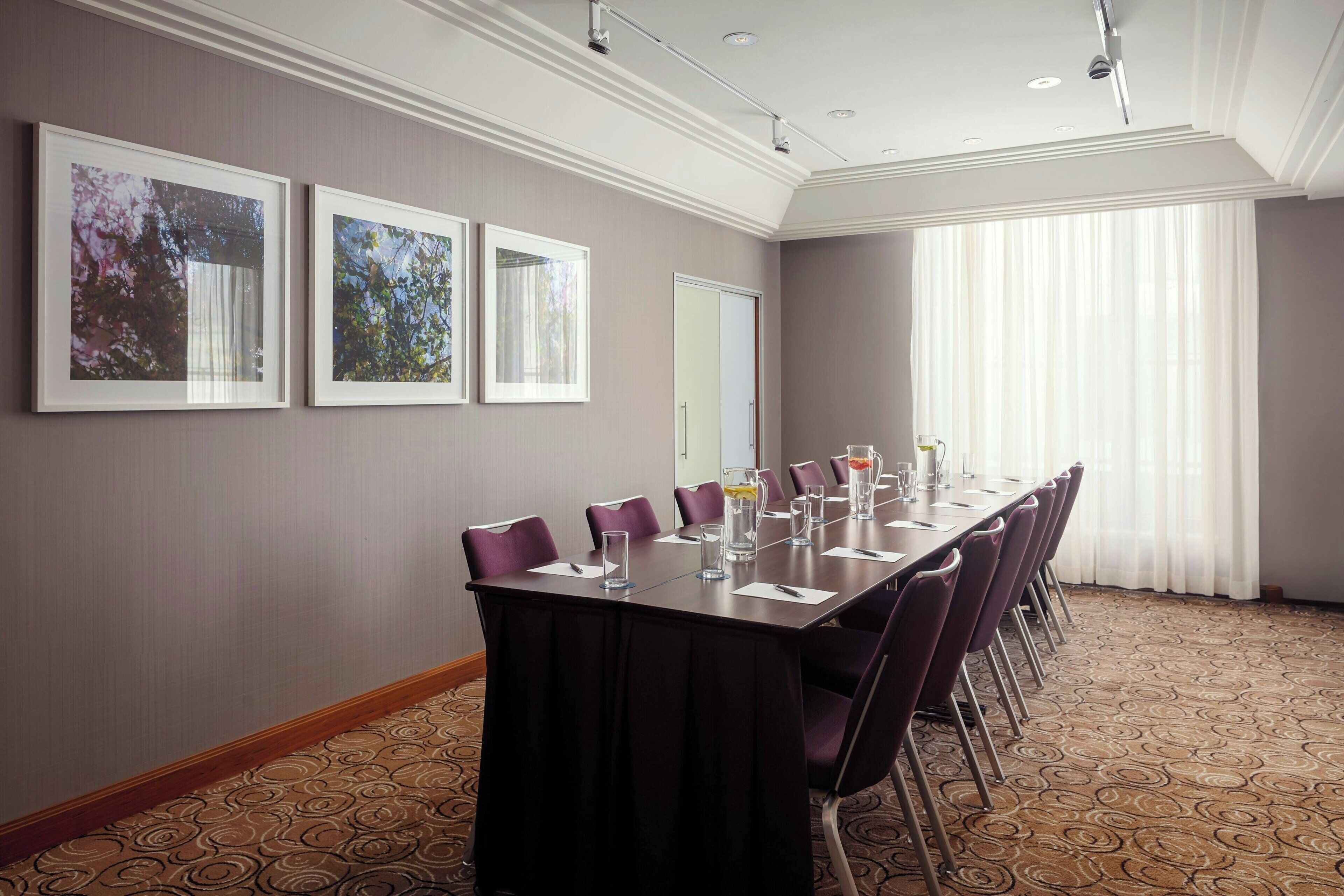 Un salón de conferencias con una larga mesa, sillas moradas, cuadros en la pared y un amplio ventanal con cortinas.