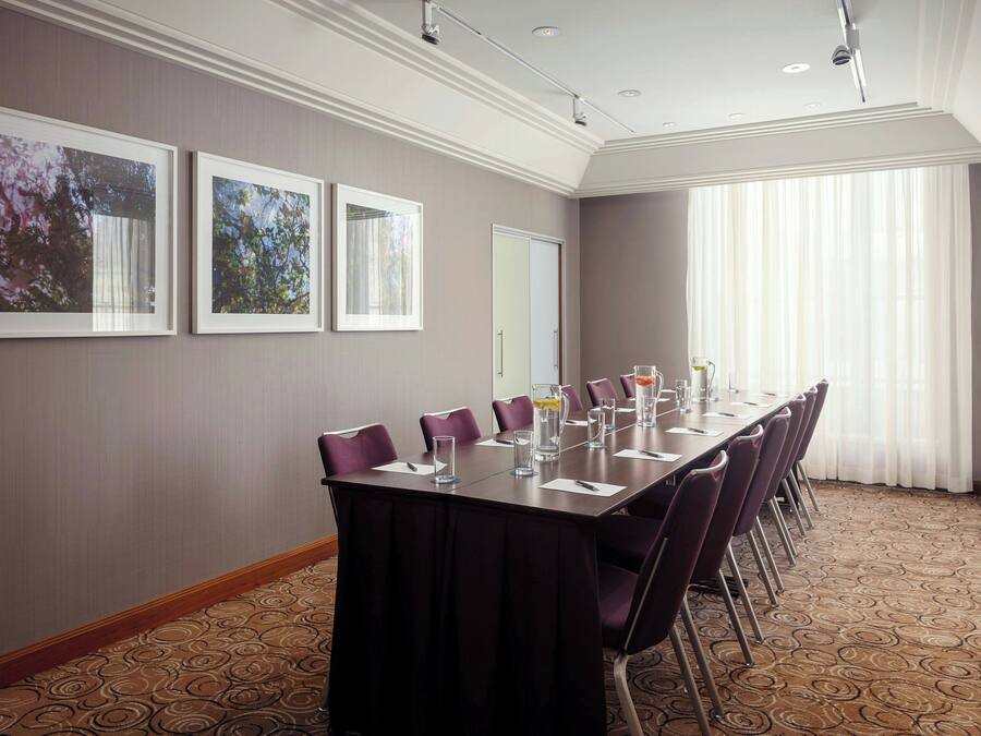 A conference room with a long table, purple chairs, framed pictures on the wall, and a large window with curtains.