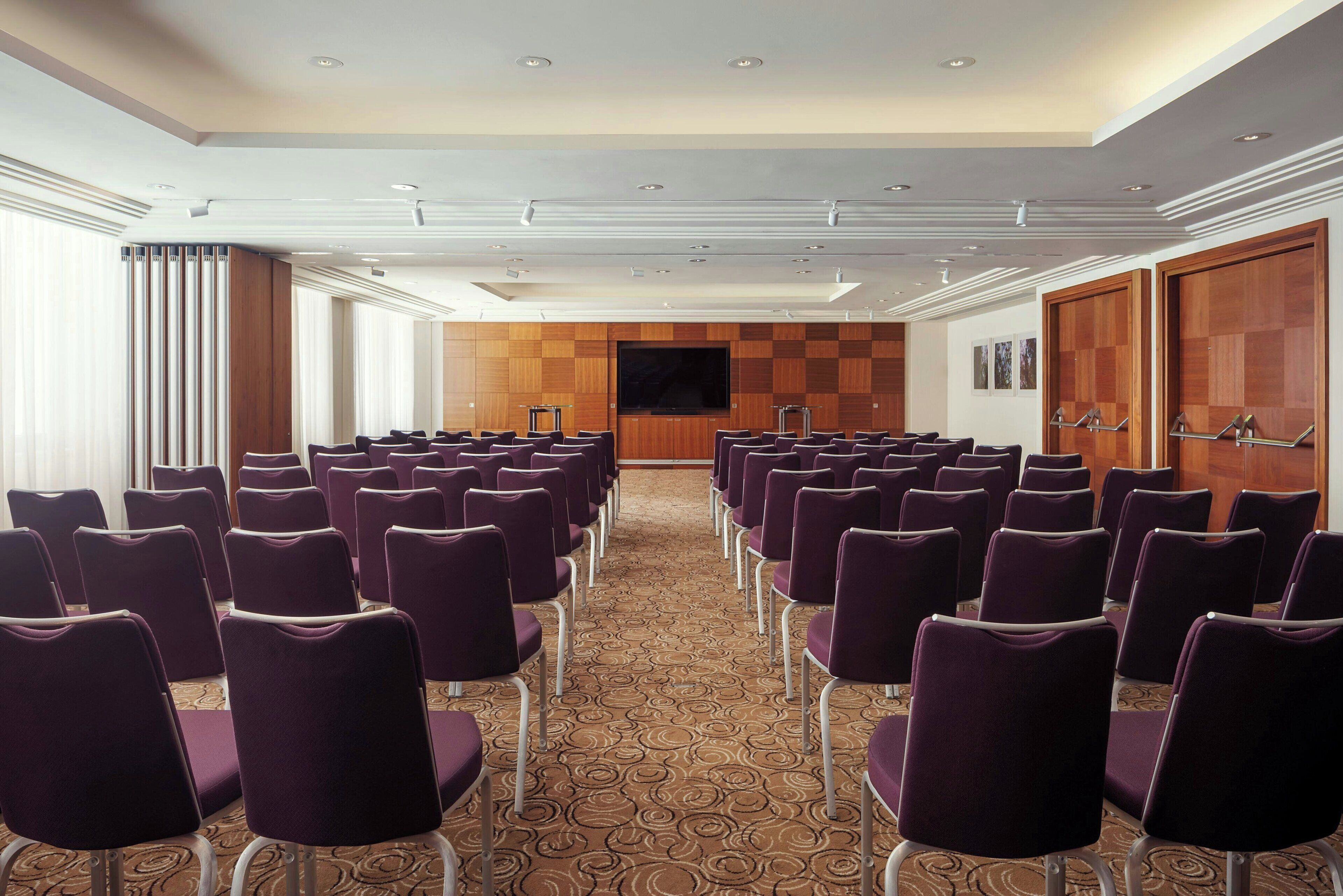 Un salón de conferencias con filas de sillas moradas, un televisor de pantalla plana grande y paneles de pared de madera.