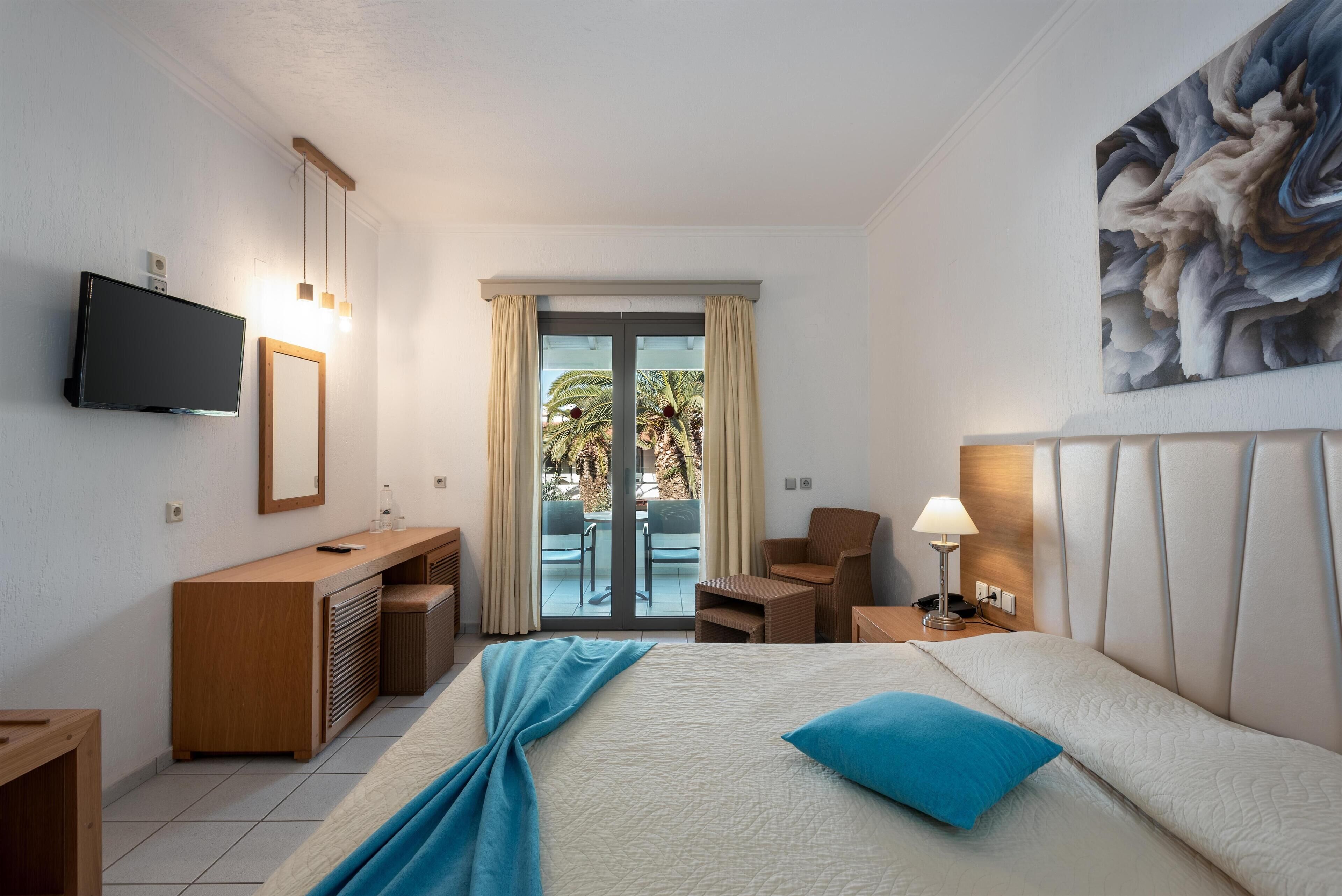 Habitación de hotel con cama, escritorio, silla, televisión y balcón con vista a palmeras.