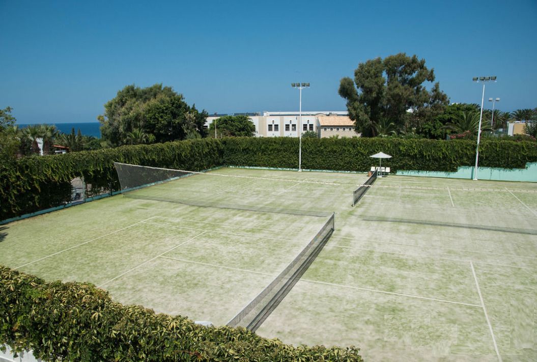 A tennis court with a net, surrounded by greenery and a building in the background.