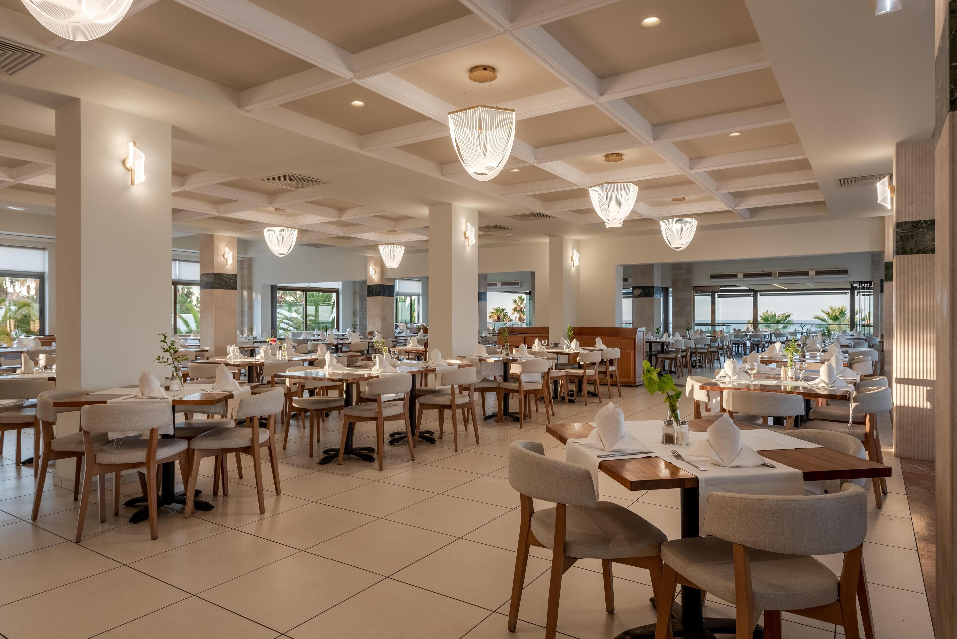 A spacious restaurant with high ceilings, chandeliers, and a modern interior design. Tables are neatly arranged with chairs, and the space is well-lit.