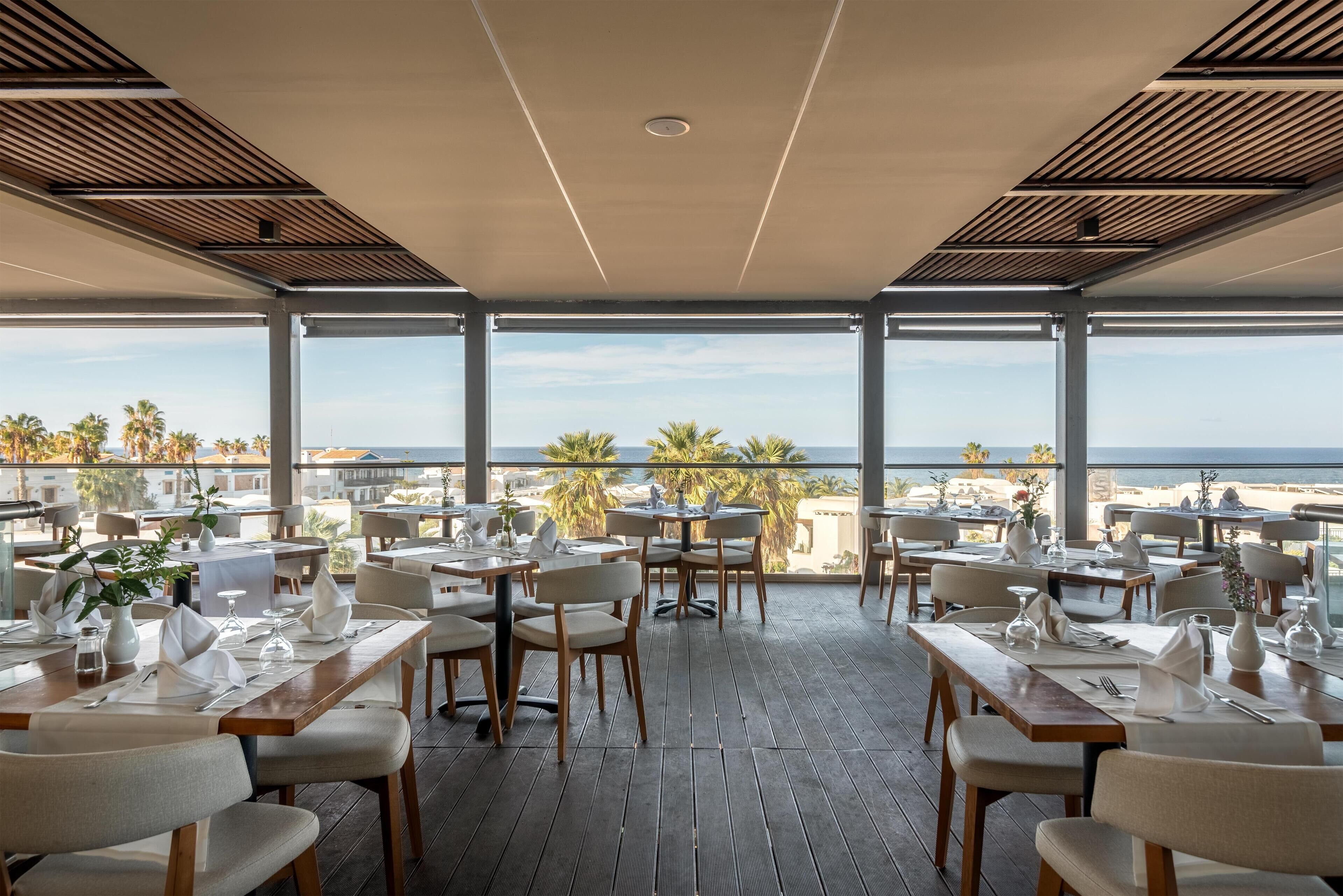 A restaurant with tables set for dining, large windows offering a view of the beach and palm trees.