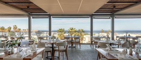 A restaurant with tables set for dining, large windows offering a view of the beach and palm trees.