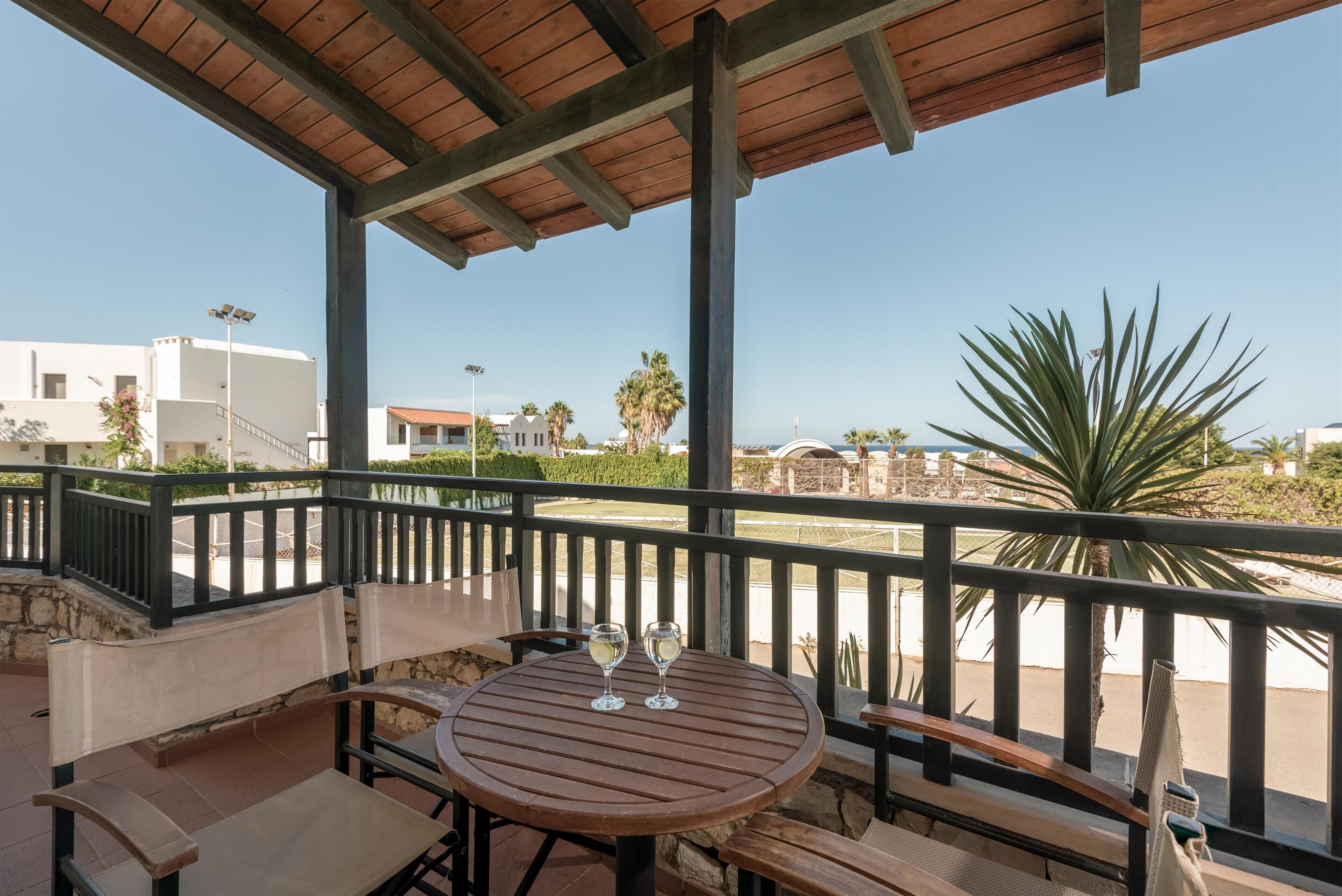 A covered outdoor seating area with a table and chairs, two glasses of white wine, and a view of the sea and buildings in the background.