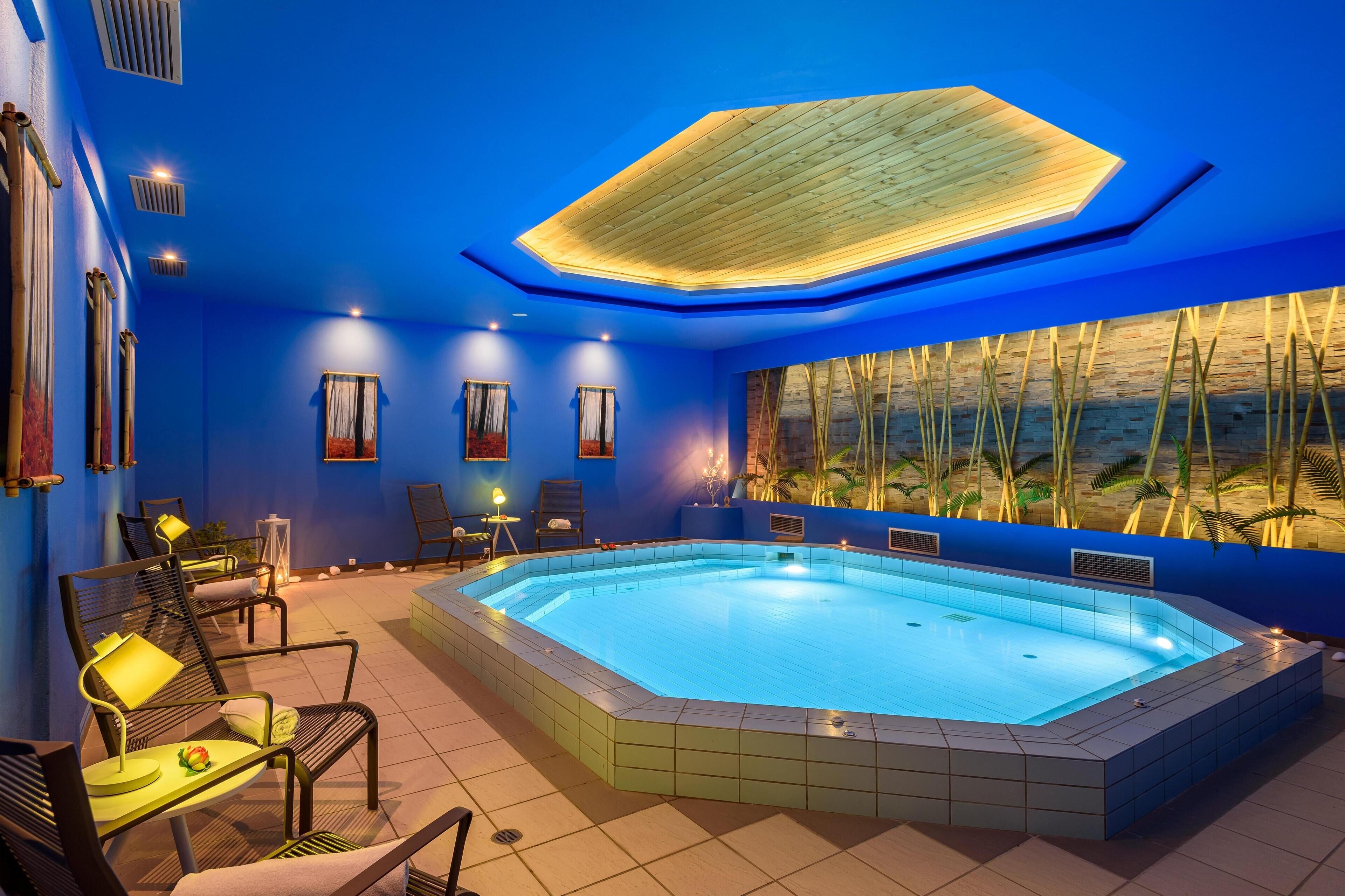 A modern indoor swimming pool with a blue-tiled floor, surrounded by lounge chairs and tables, and a large window with a view of palm trees.