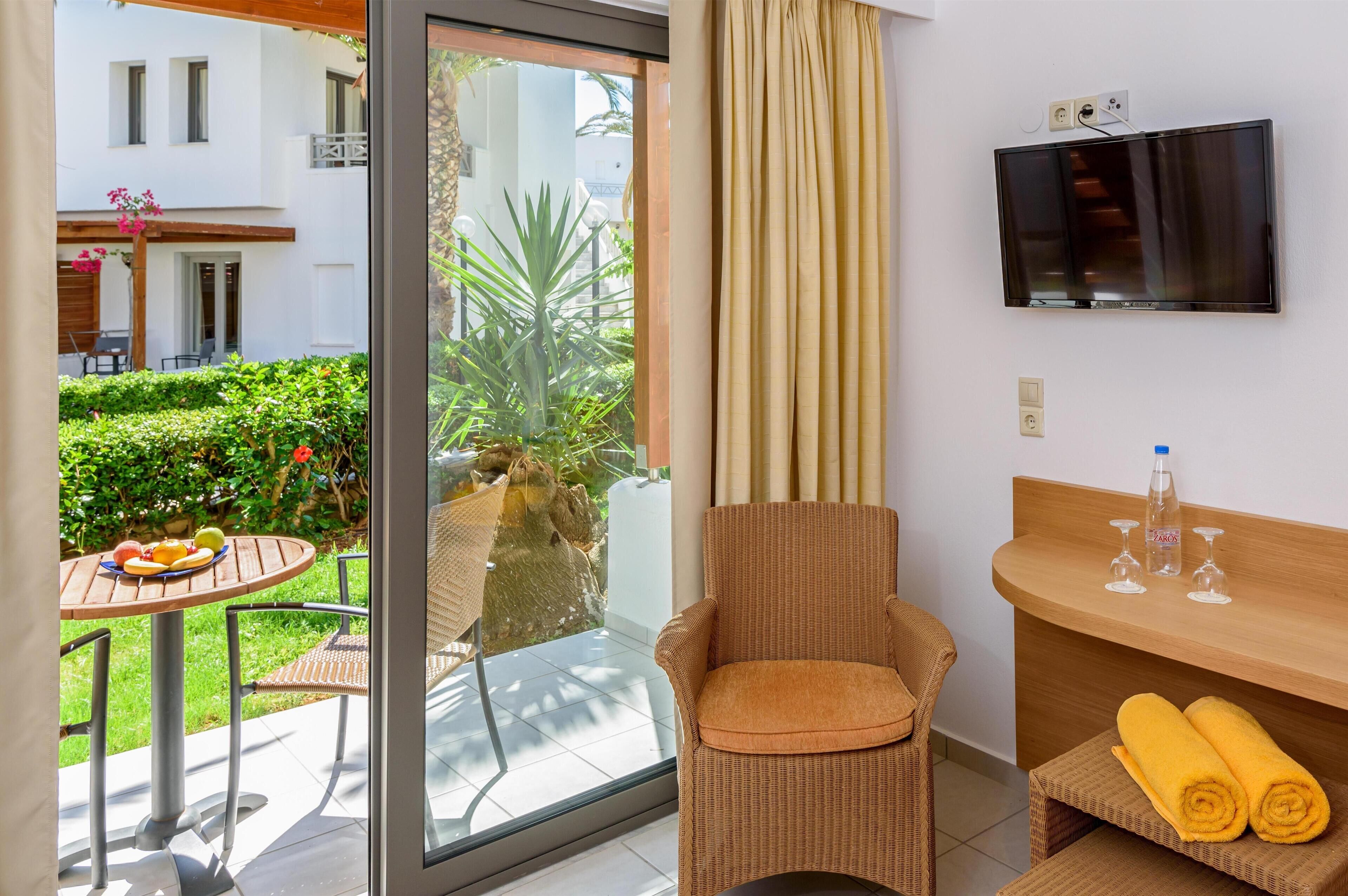 A hotel room with a glass door leading to a balcony, a wicker chair, a small table with fruits, a TV mounted on the wall, and a wooden side table with towels and glasses.