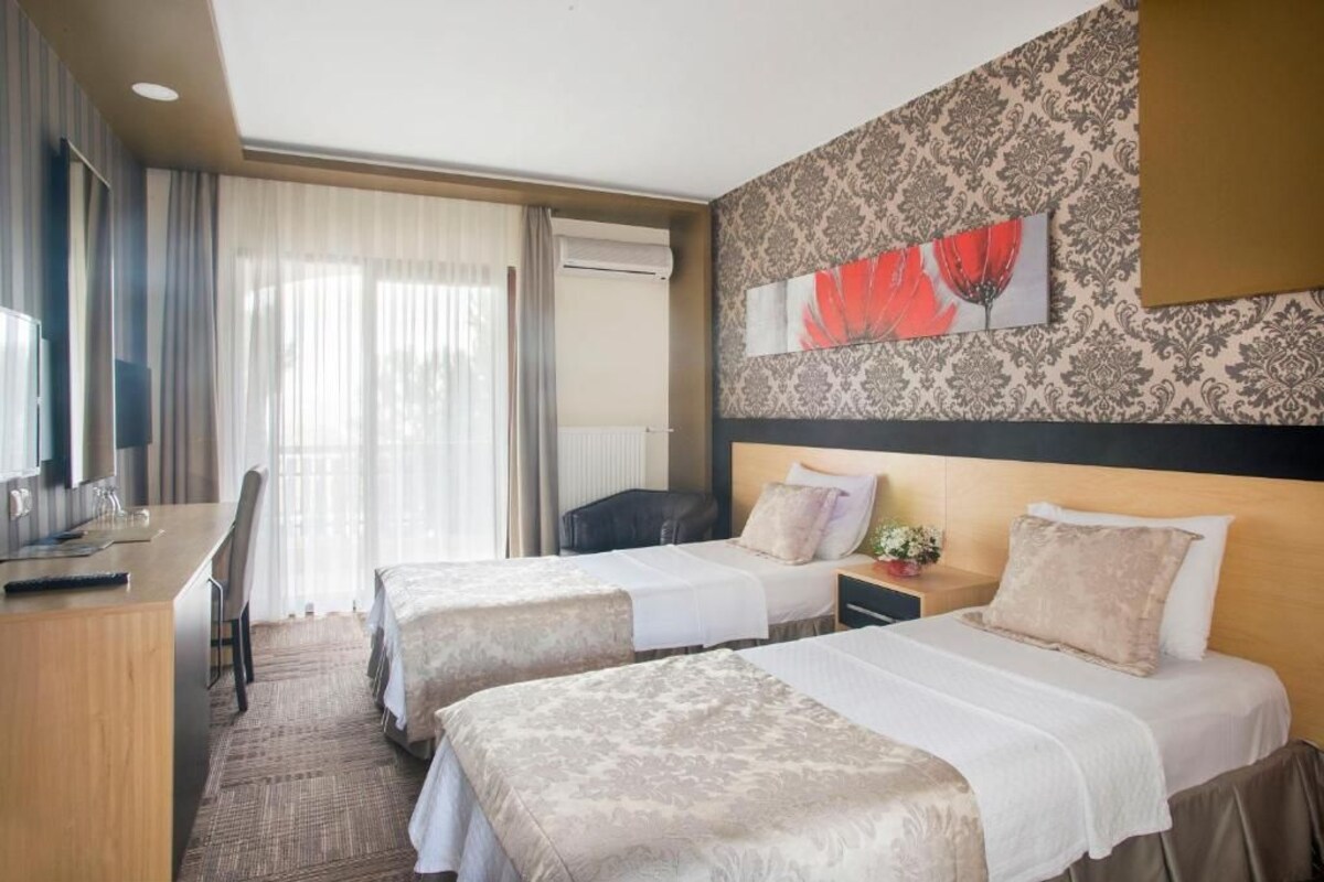 A hotel room with two beds, a desk, and a chair. There is a window with curtains, a wall with a floral pattern, and a painting of red flowers.
