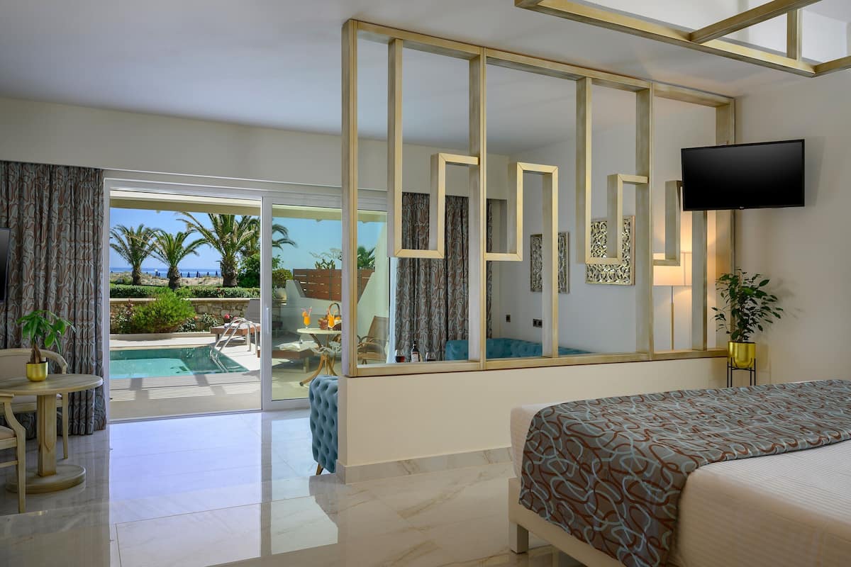 A hotel room with a bed, a TV, a large window with a view of a pool and palm trees, and a dining area with a table and chairs.