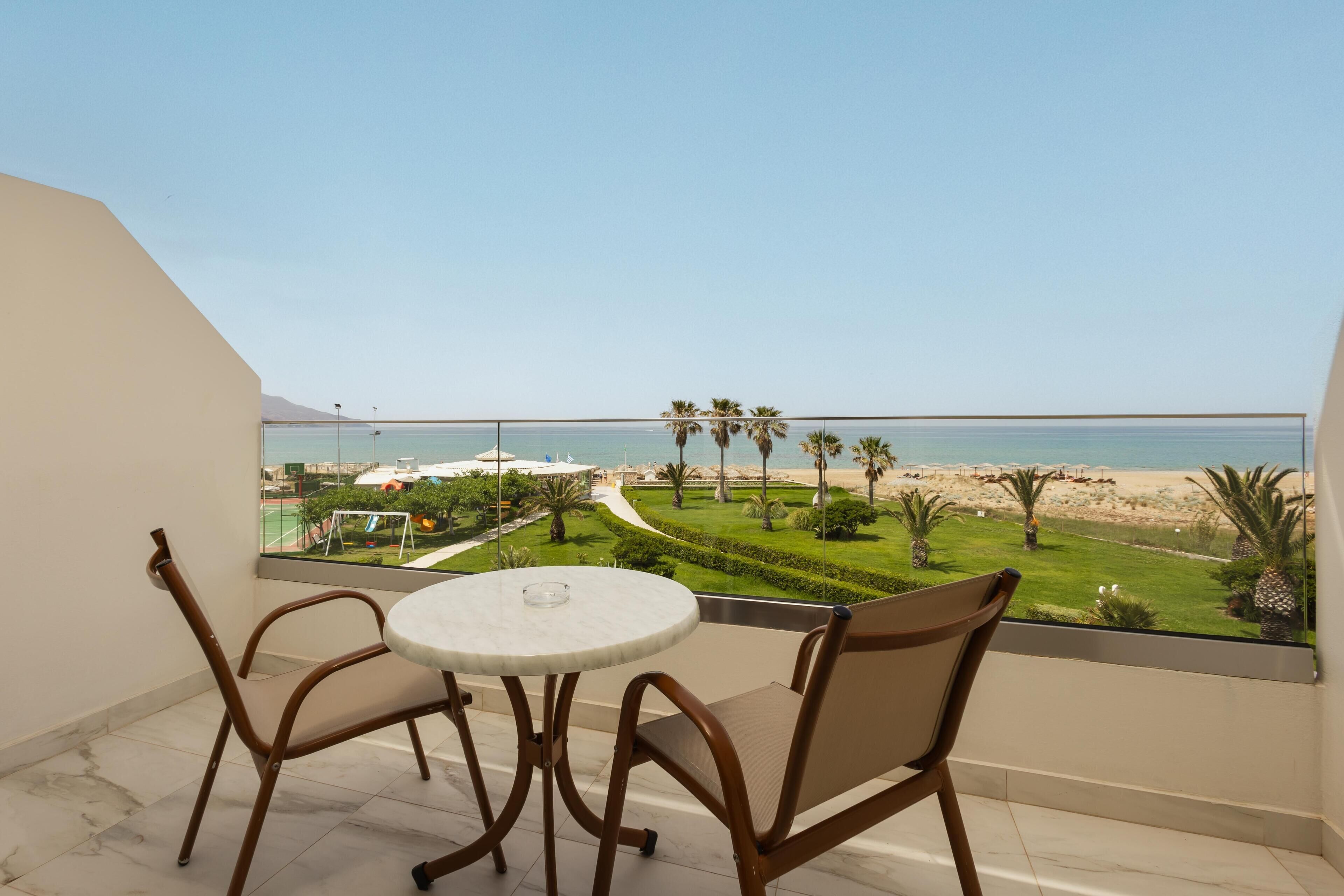 Un balcon avec une table et des chaises, donnant sur une plage bordée de palmiers et offrant une vue sur un ciel dégagé.
