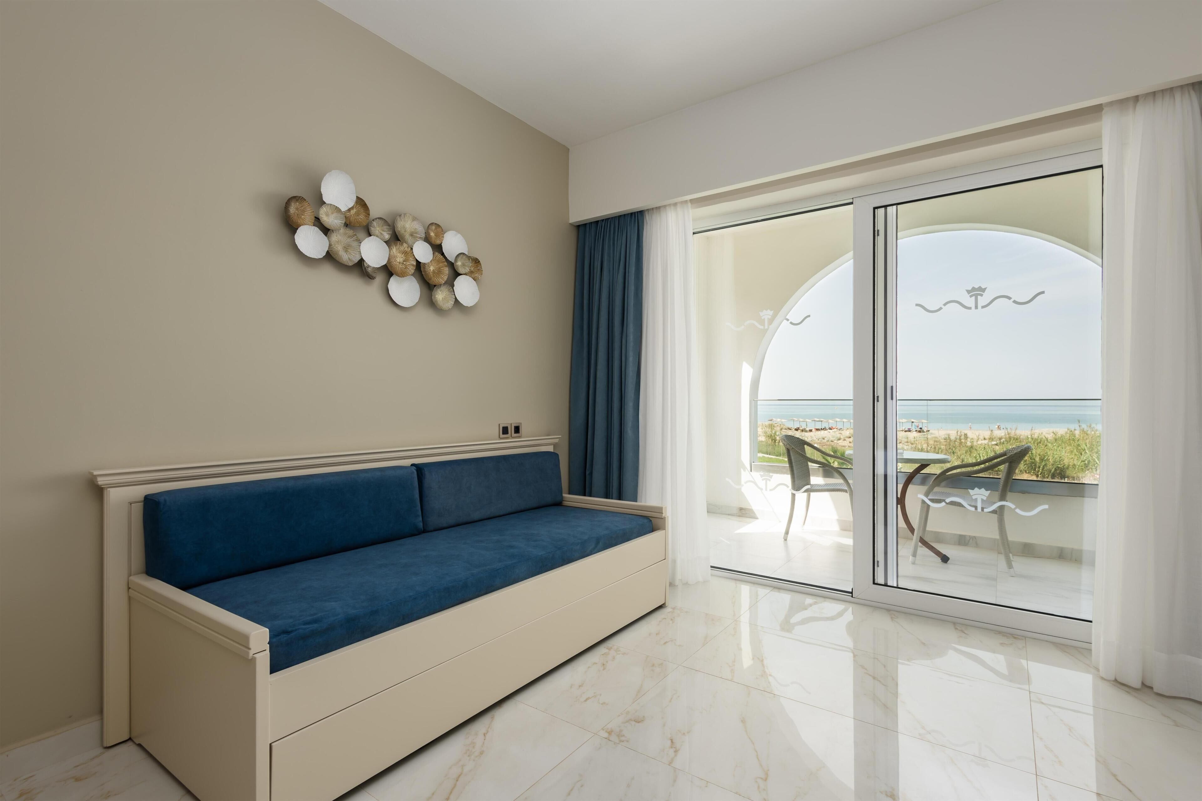 Un salon moderne avec un canapé bleu, un élément décoratif mural et une vue sur la plage, visible à travers de grandes portes vitrées.