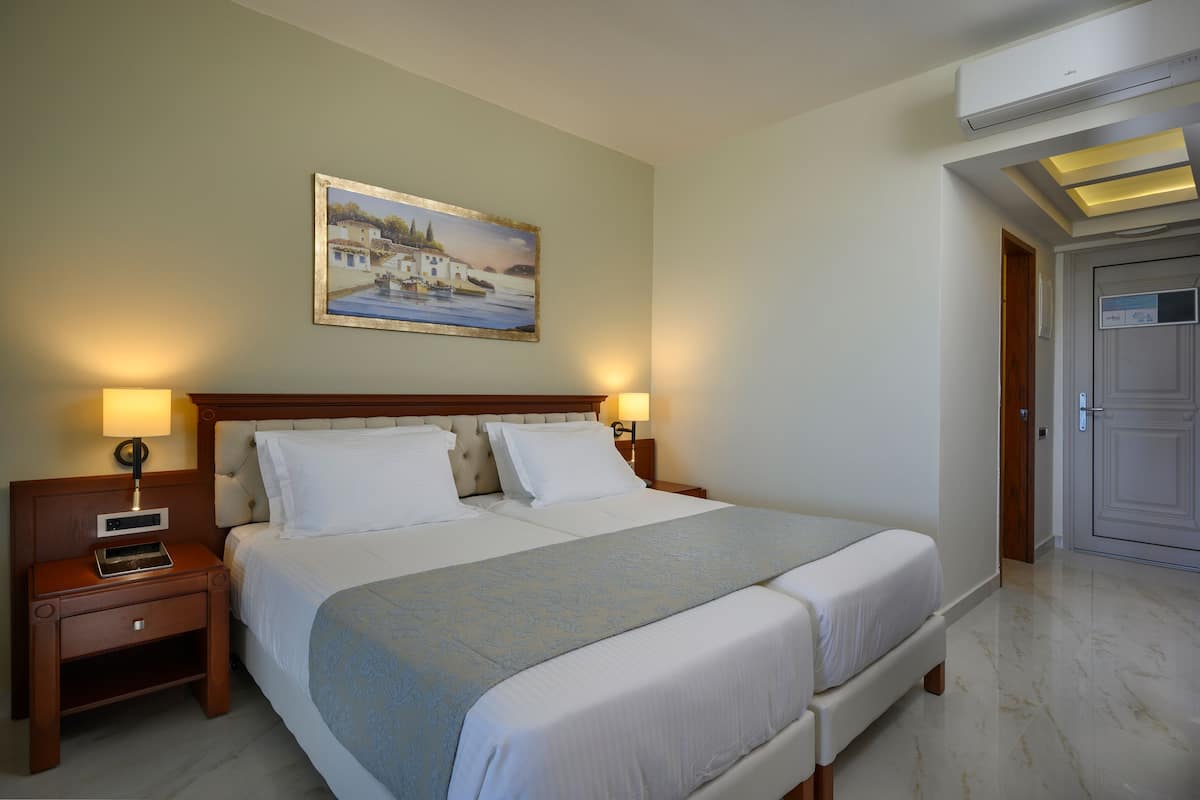 A hotel room with a large bed, two bedside tables with lamps, a painting on the wall, and an air conditioning unit.