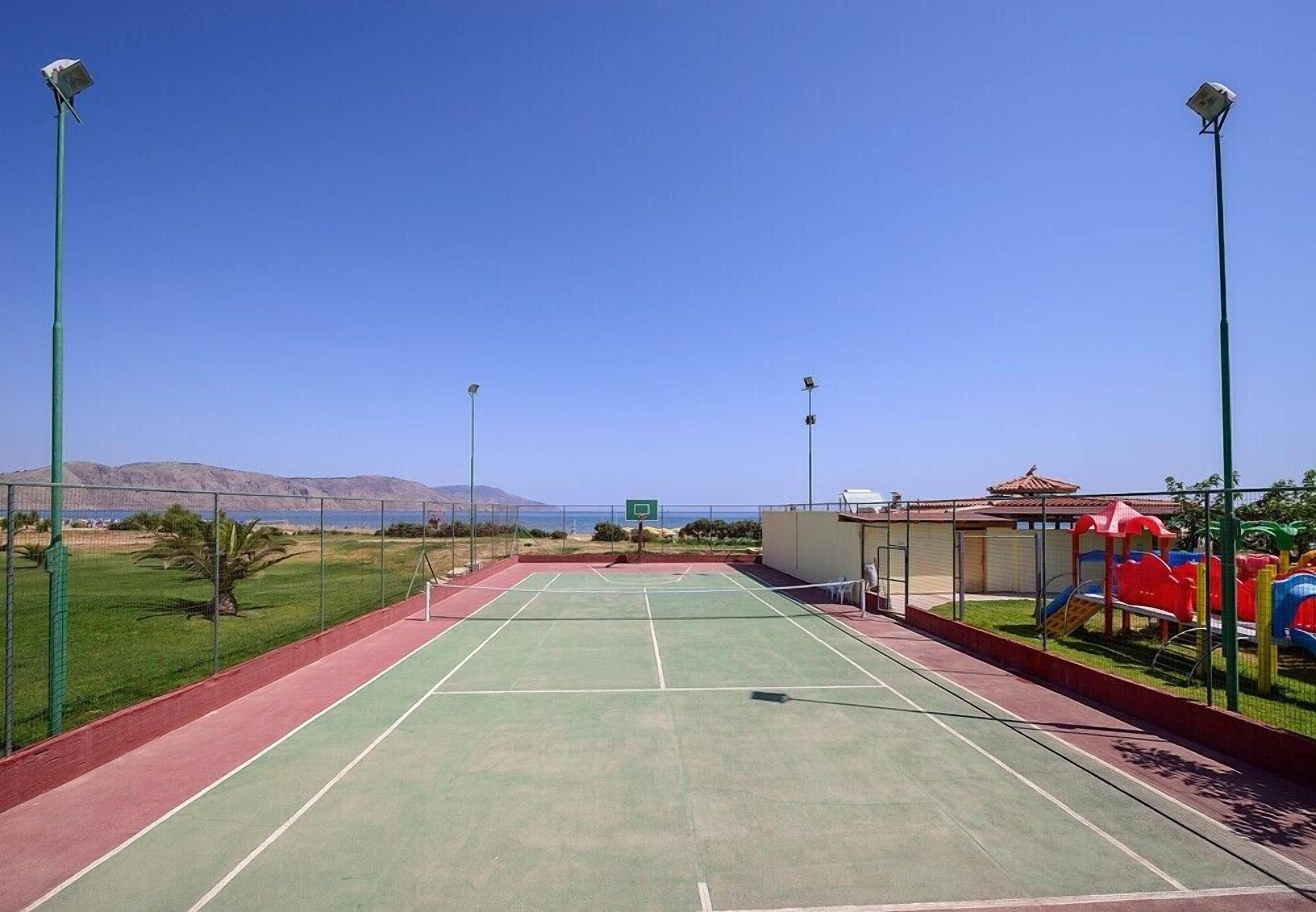 Una cancha de tenis al aire libre con superficie verde, rodeada por una valla y equipada con iluminación.