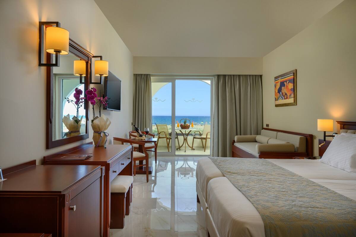 A hotel room with a bed, a desk, a chair, a TV, a large window with a view of the beach, and a painting on the wall.