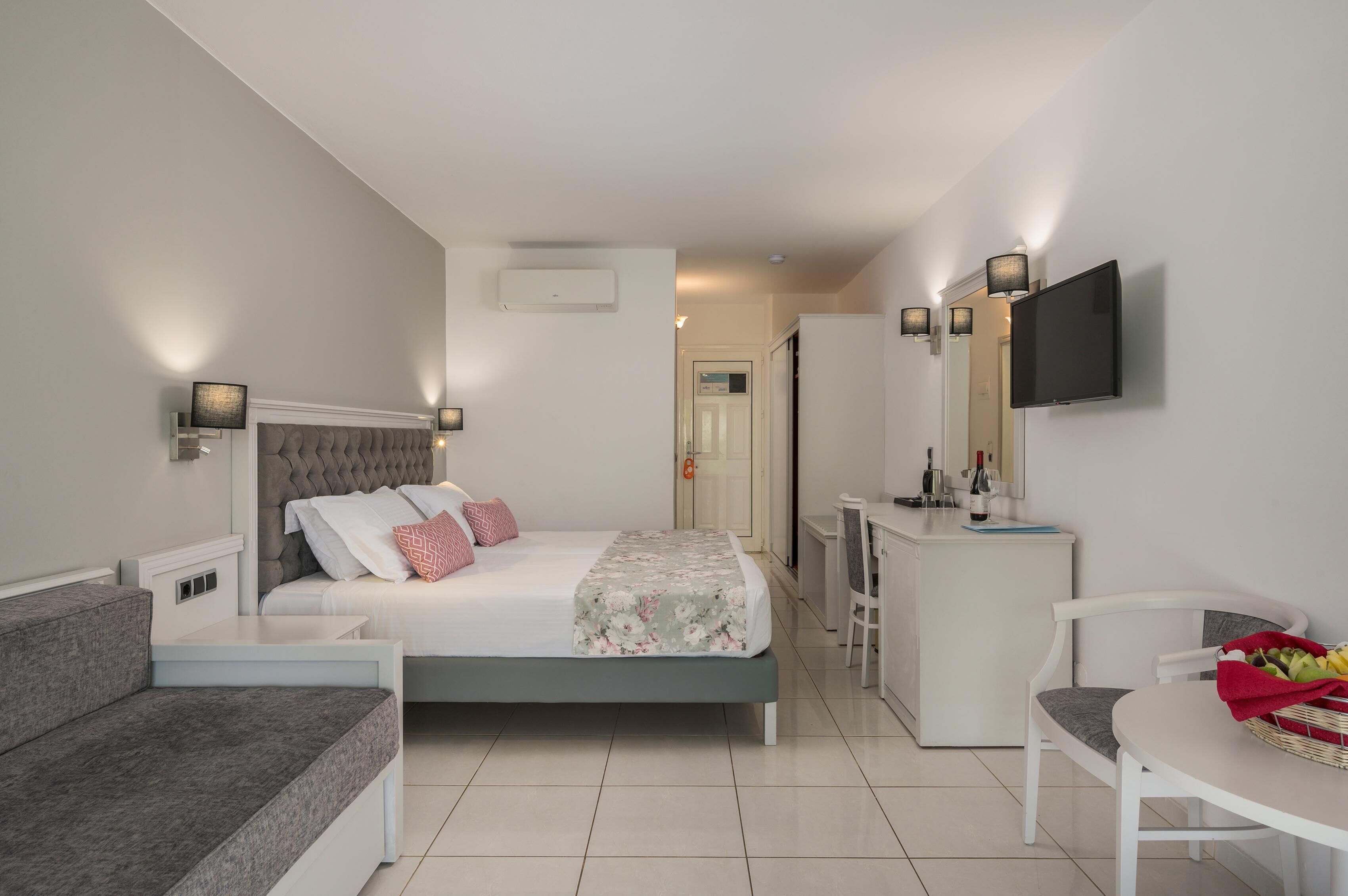 Un dormitorio moderno con cama, mesitas de noche, un escritorio, una silla, televisor y una pequeña kitchenette.