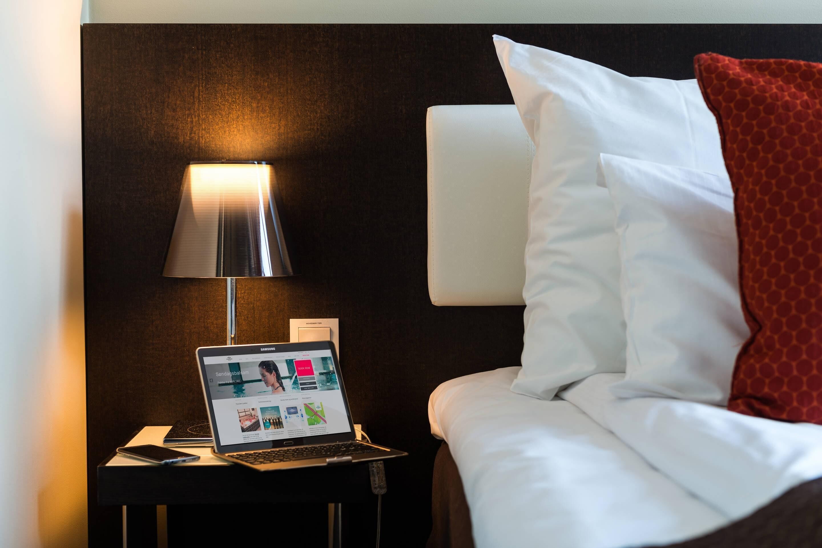 Una habitación de hotel con cama, mesita de noche con una computadora portátil, lámpara y teléfono.