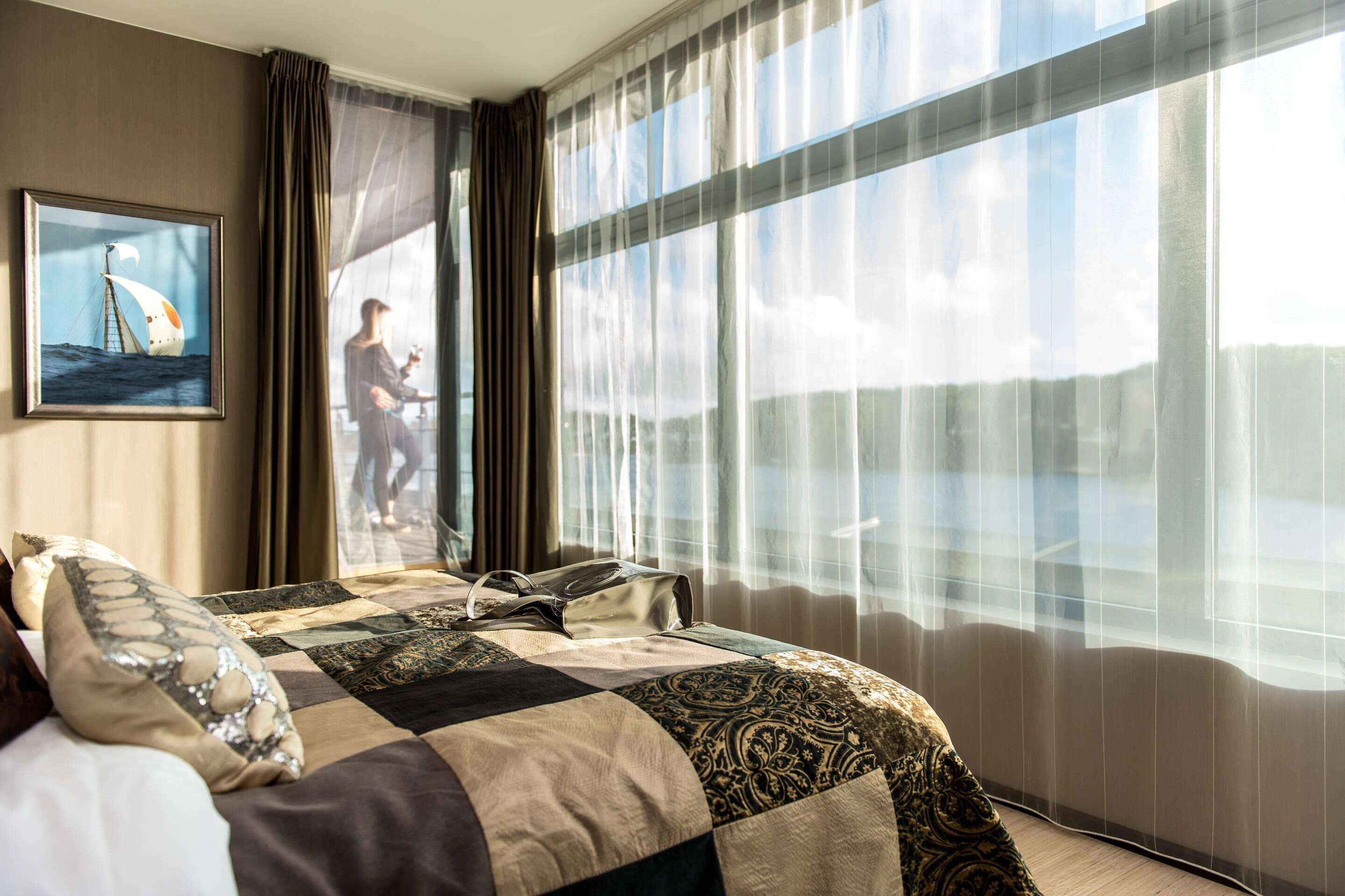 Una habitación de hotel con una cama grande, vista al mar a través de cortinas traslúcidas y un cuadro con una imagen de un velero en la pared.