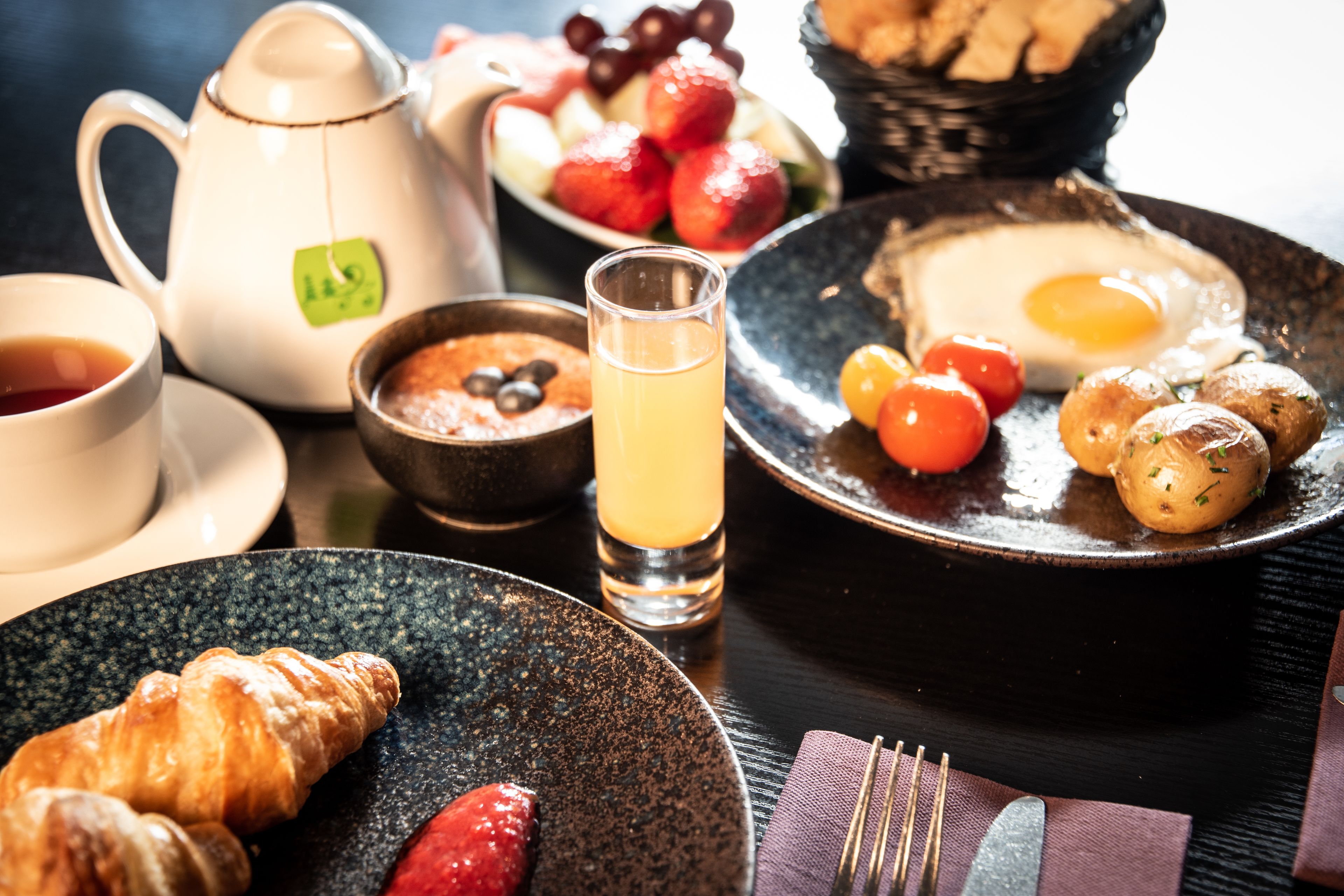 Une table mise pour le petit-déjeuner avec une théière, des tasses, un verre de jus, des croissants, des fraises et une assiette contenant des œufs et des tomates.