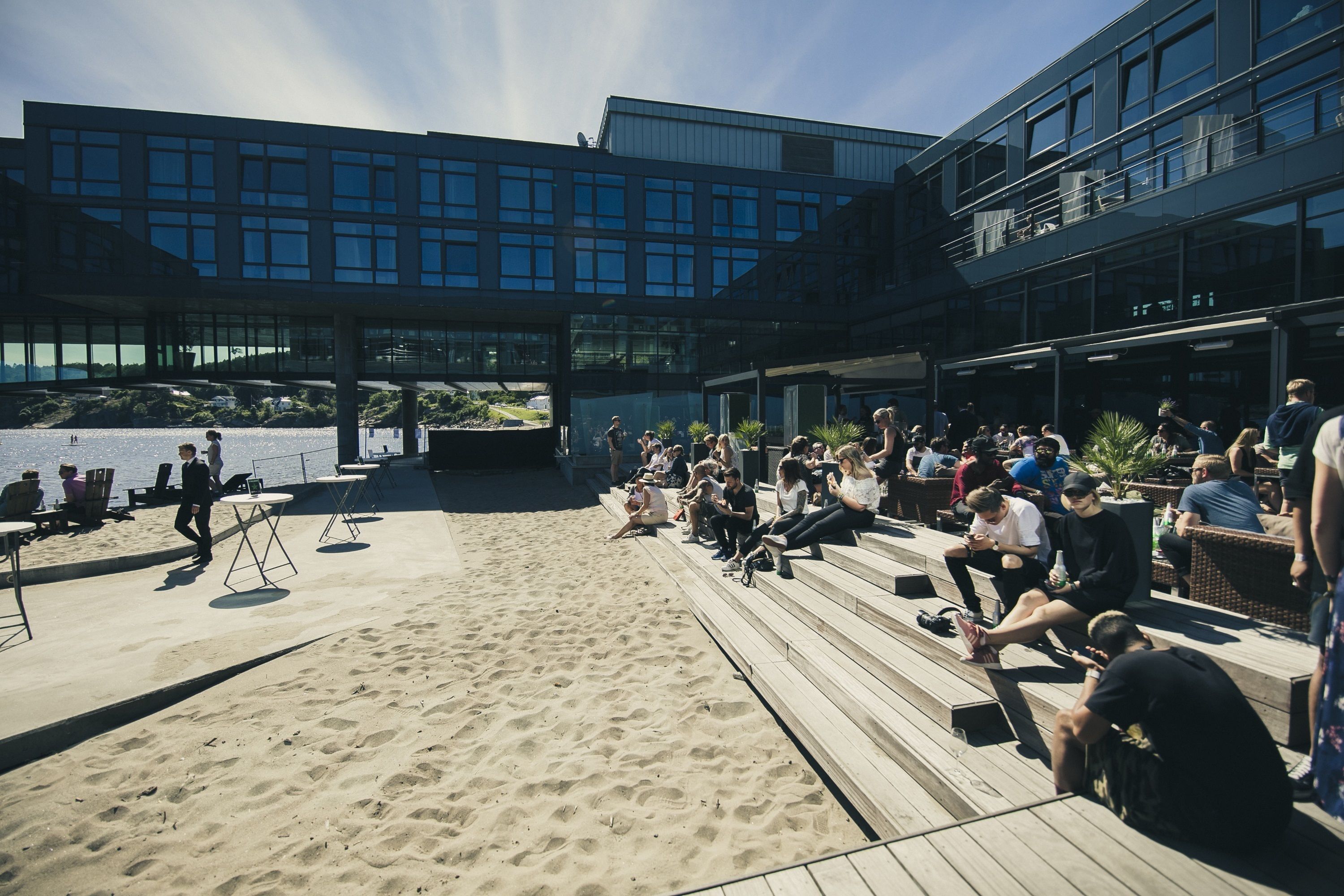 An outdoor seating area with a sandy floor and wooden steps, overlooking a body of water and a modern building with large windows.
