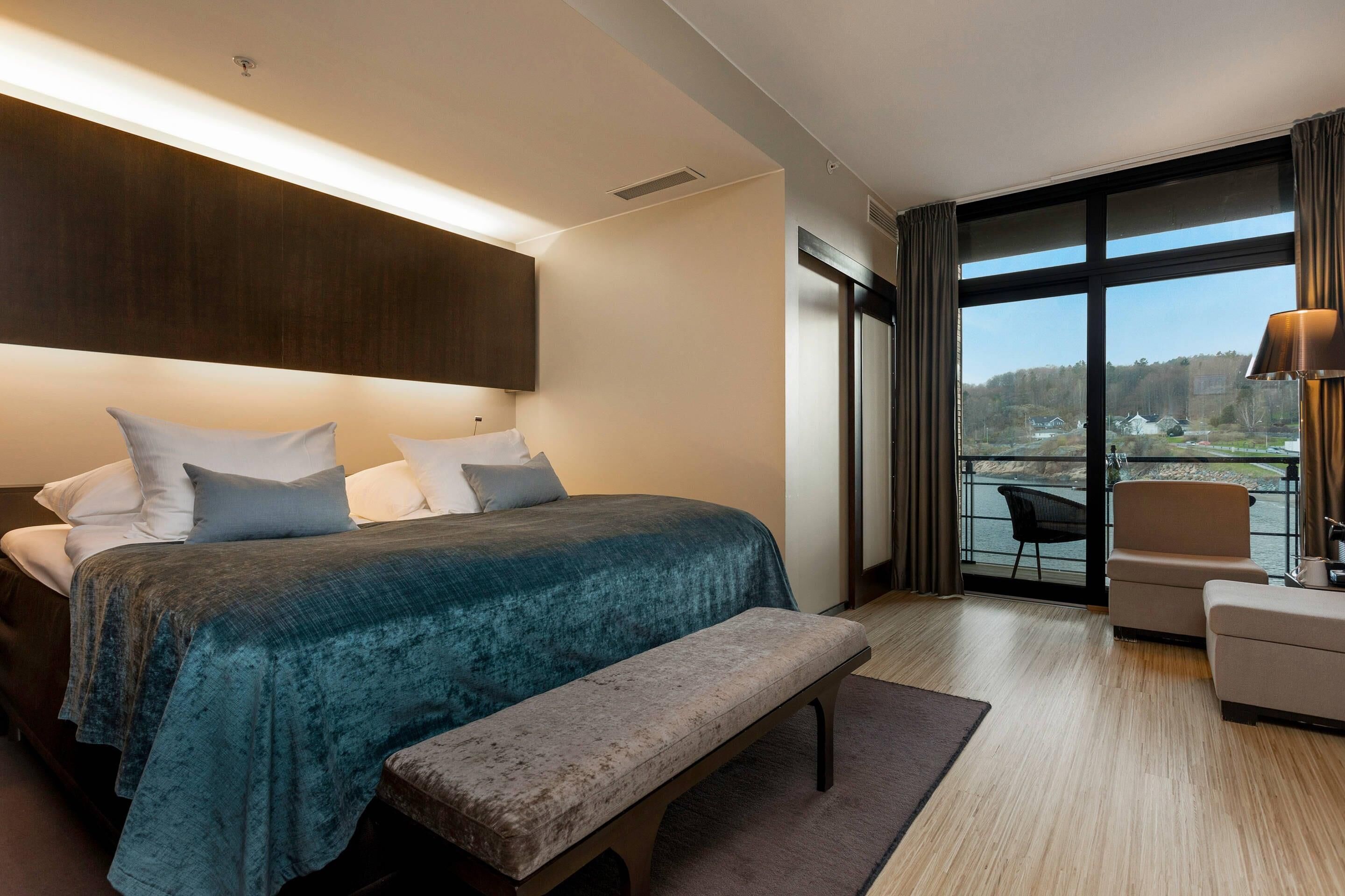 A modern hotel room with a large bed, a bench, a seating area with a chair, and a view through a large window.