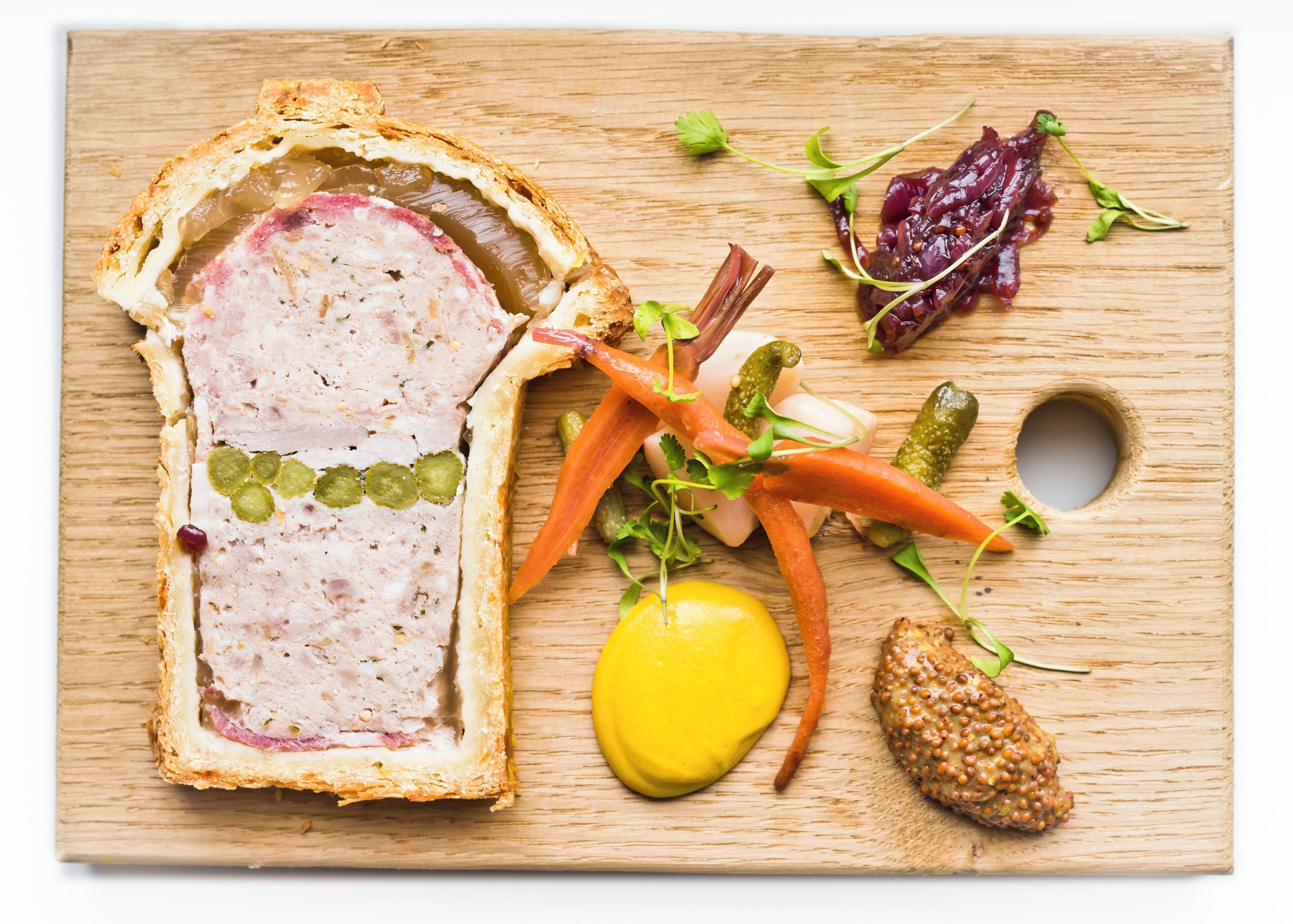 Un trozo de pastel de carne con costra, adornado con vegetales y un gajo de limón sobre una tabla de madera.