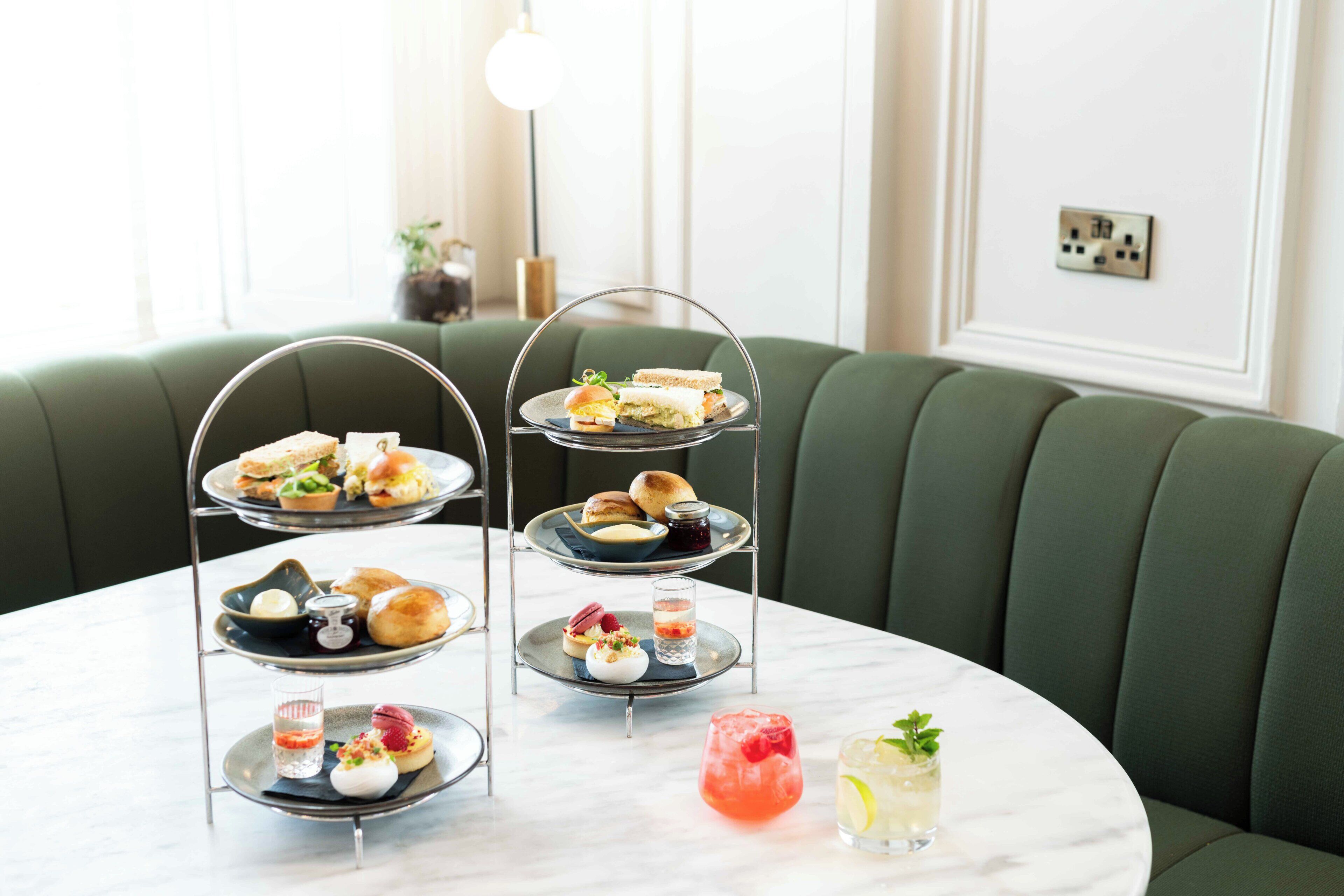 A three-tiered dessert stand with various pastries and a drink on a marble table in a green upholstered seating area.