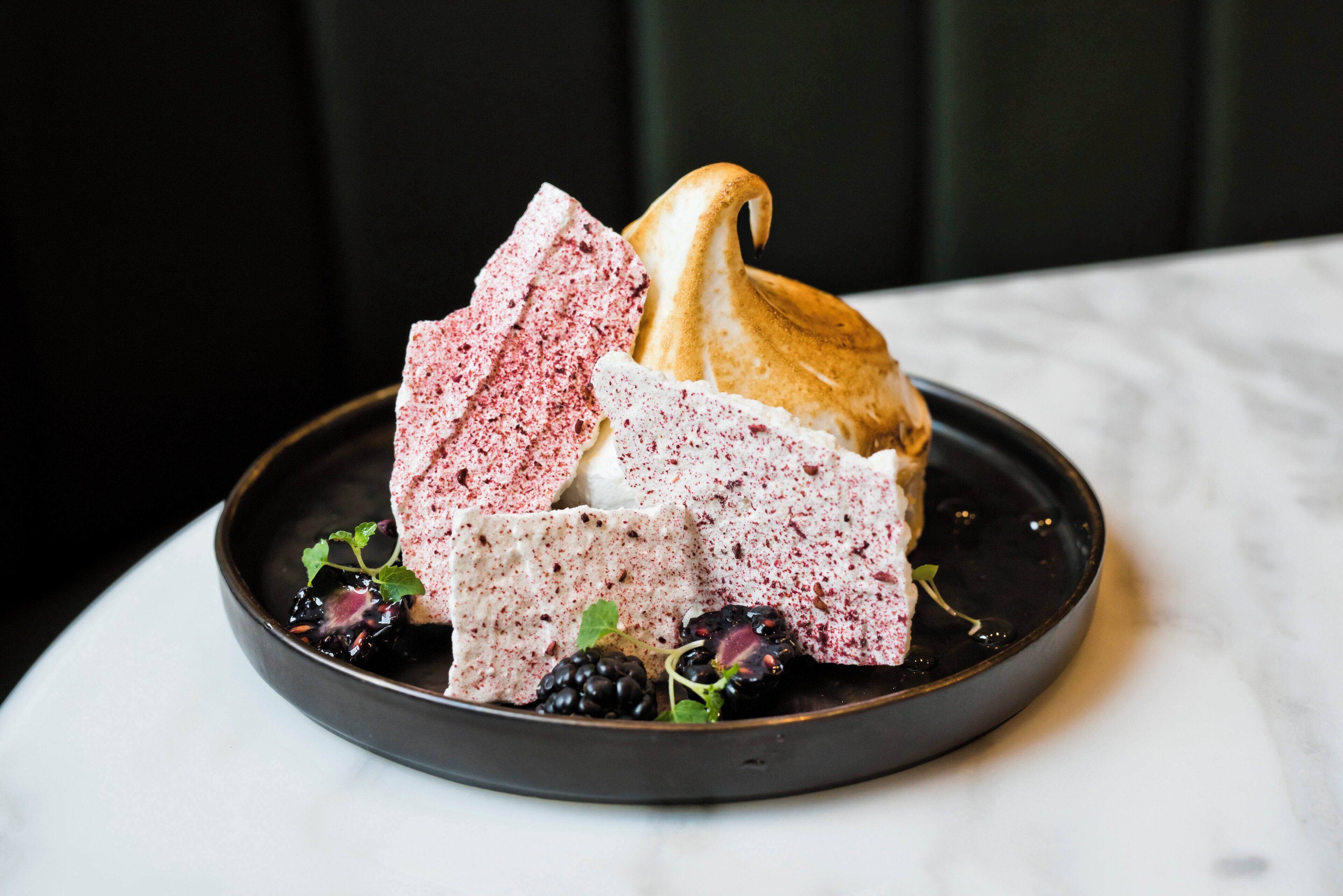 A dessert plate with a meringue, a pink cake, and blackberries on a dark plate.