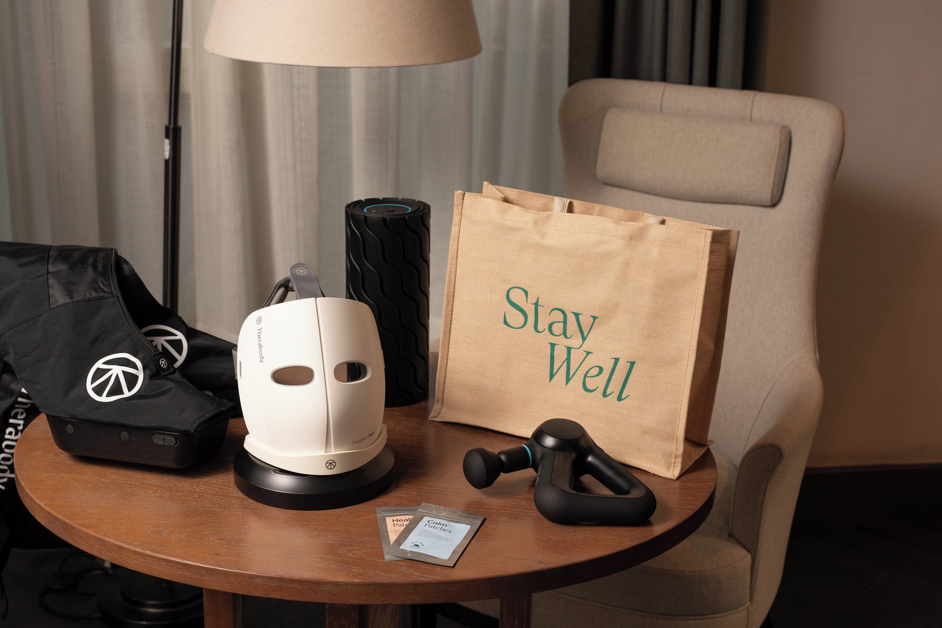 A table with a massage gun, a white massage chair, a black massage chair, a brown paper bag with "Stay Well" text, and a black bag with a peace symbol.