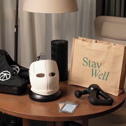A table with a massage gun, a white massage chair, a black massage chair, a brown paper bag with "Stay Well" text, and a black bag with a peace symbol.