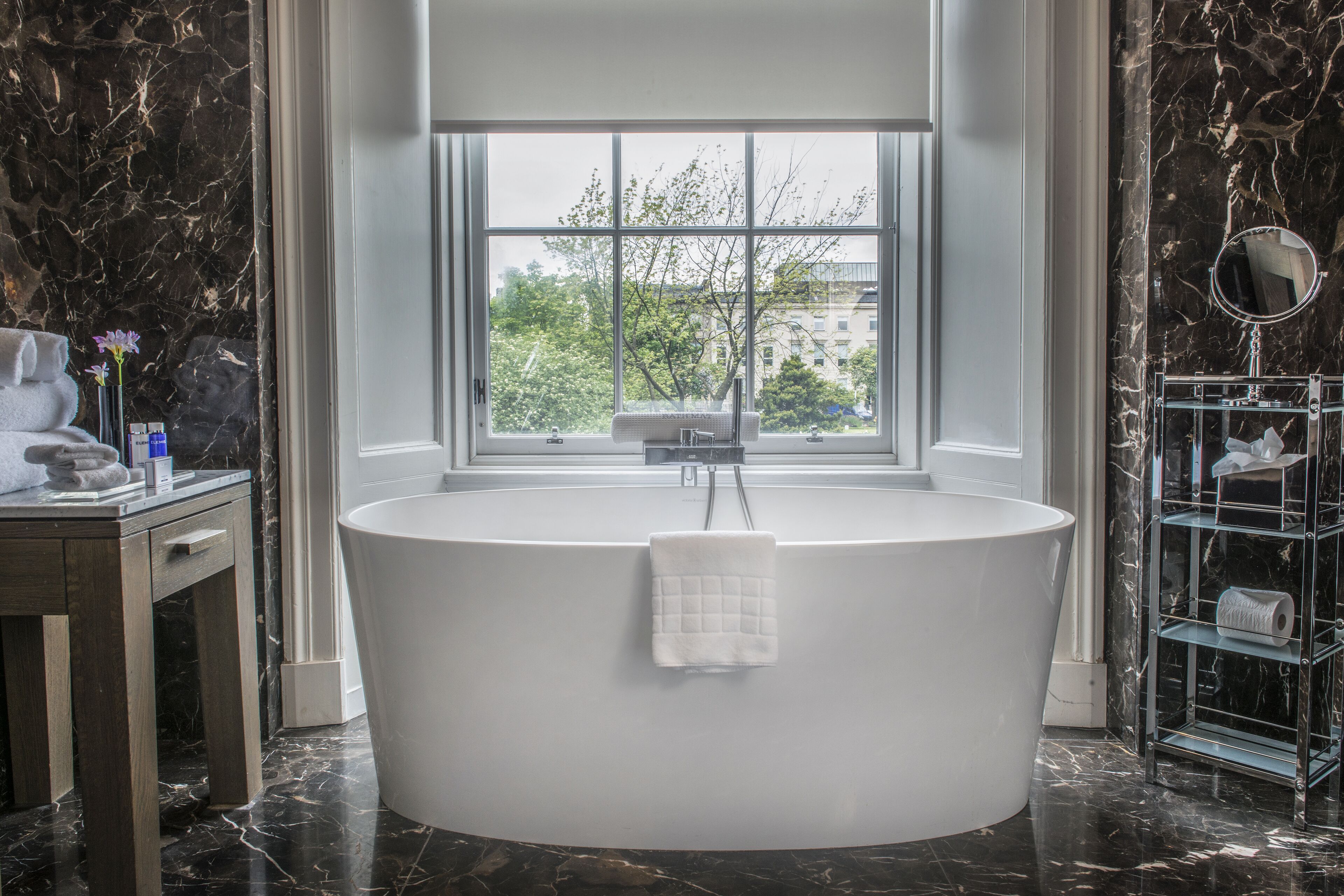Un baño moderno con una bañera independiente, una ventana que ofrece vistas a árboles y edificios, suelo de mármol y pared de mármol.