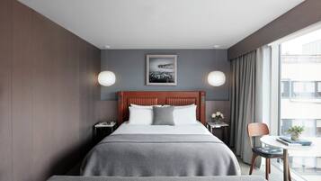 A hotel room with a large bed, a bedside table with a lamp, a framed picture on the wall, and a view of the city through the window.