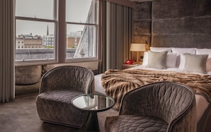 A hotel room with a bed, two armchairs, a small table, a nightstand with a lamp, and a view of buildings through large windows.