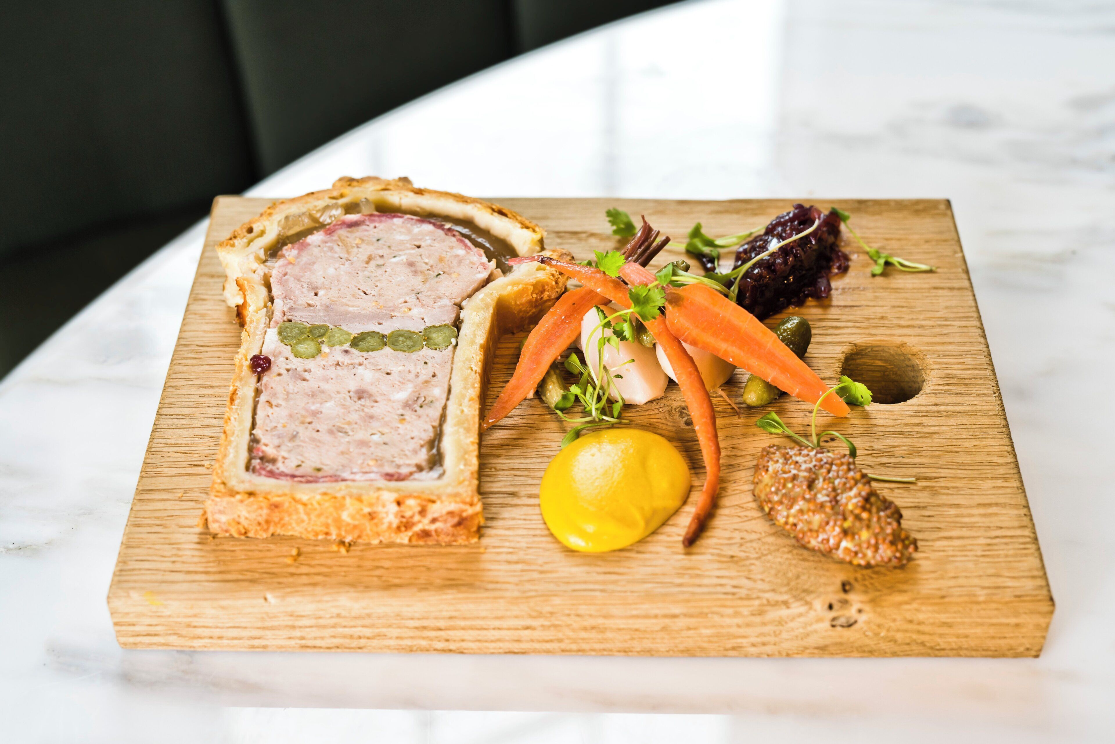 A wooden board with a slice of meatloaf, a lemon, and some vegetables.