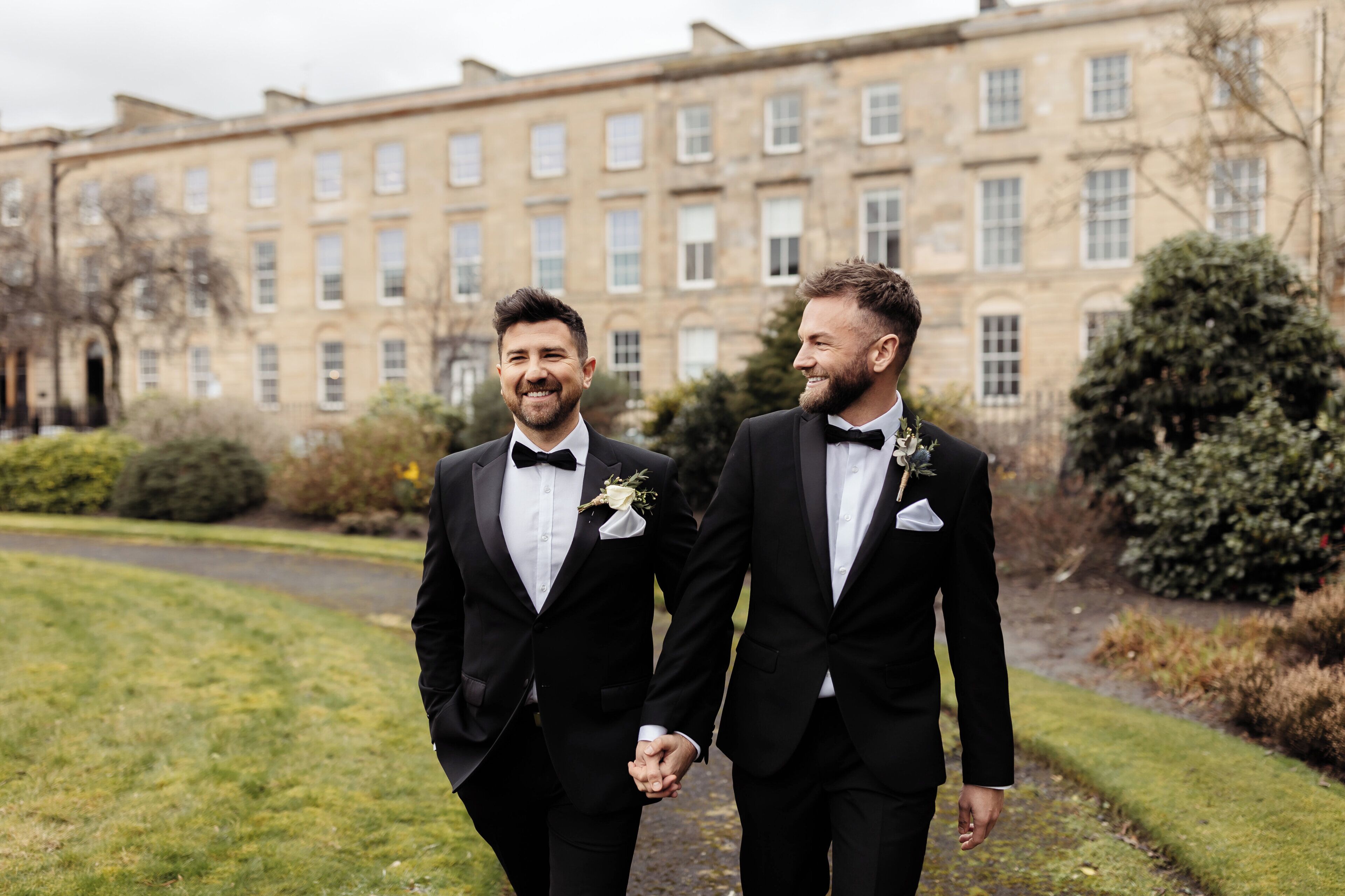 Two men in black suits holding hands in front of a large building with multiple windows.