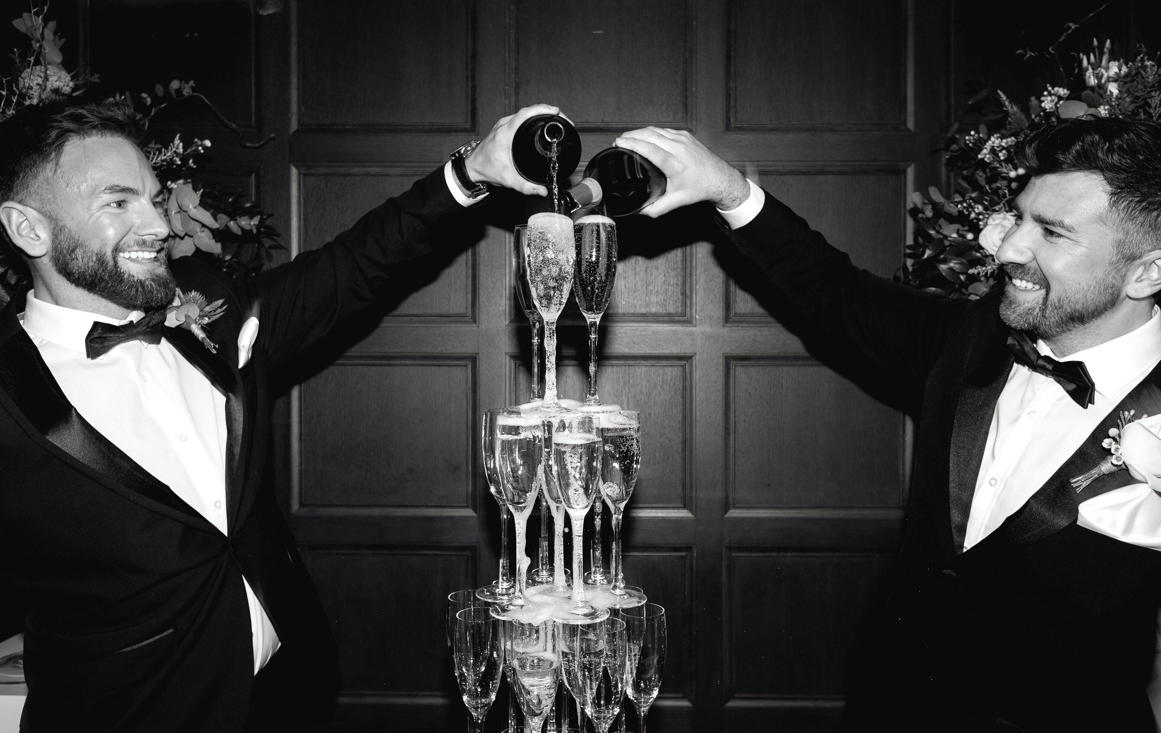Two men in formal attire holding a champagne bottle above a stack of champagne glasses.