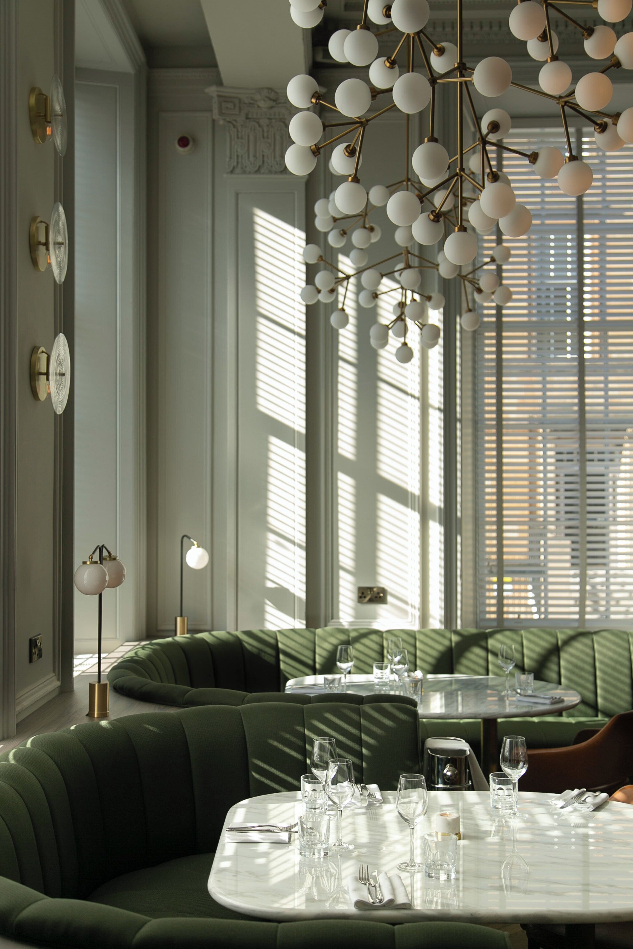 Un espace repas avec des banquettes recouvertes d’un tissu vert, une table ronde dressée pour le dîner et un lustre moderne doté de luminaires sphériques.