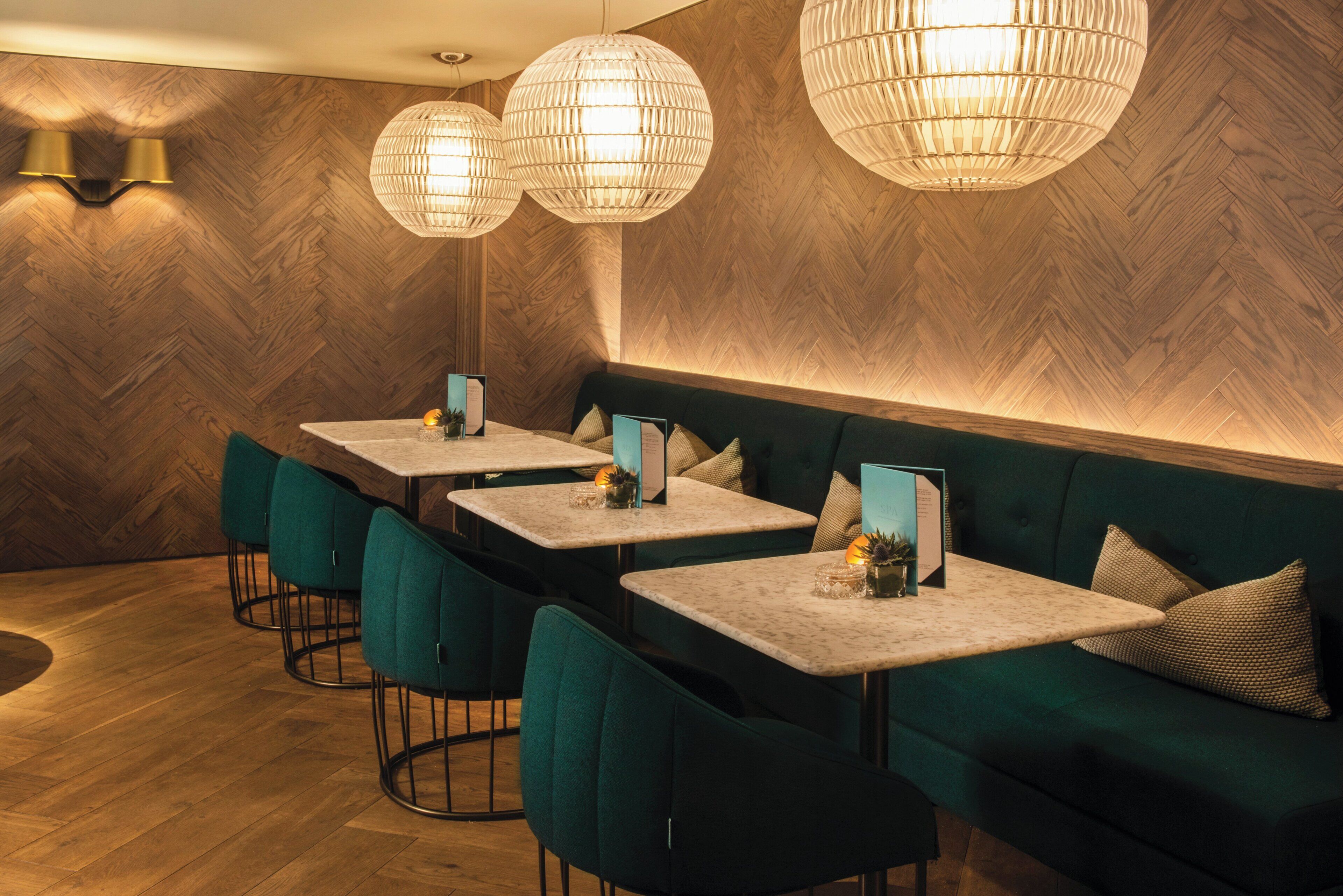 A restaurant interior with herringbone wood flooring, green upholstered seating, and pendant lighting.