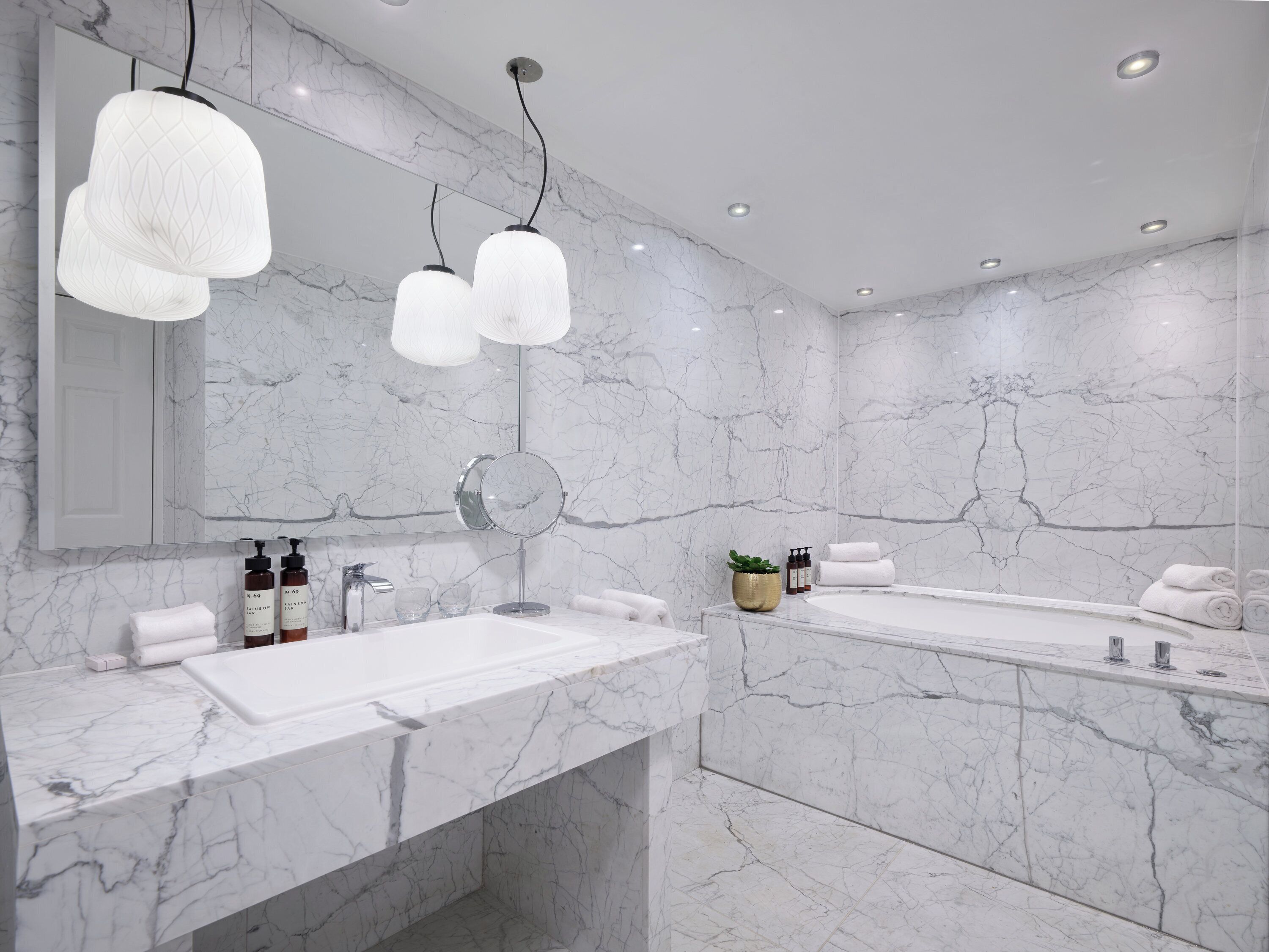 A modern bathroom with marble walls, a large mirror, two sinks, and pendant lights.