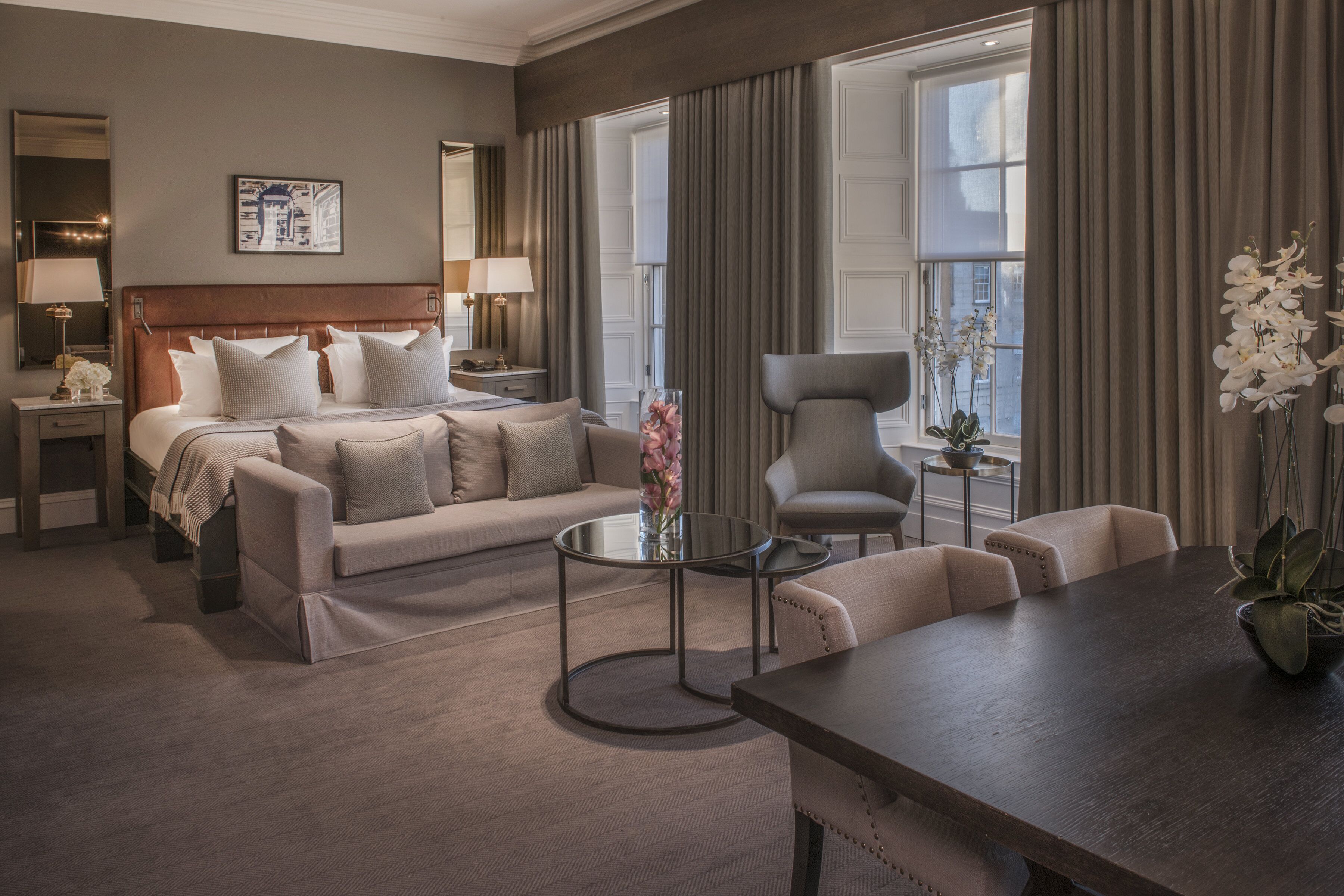 Habitación de hotel con cama, sofá, mesa de comedor y una silla. Hay una ventana con cortinas, una lámpara y un jarrón con flores.