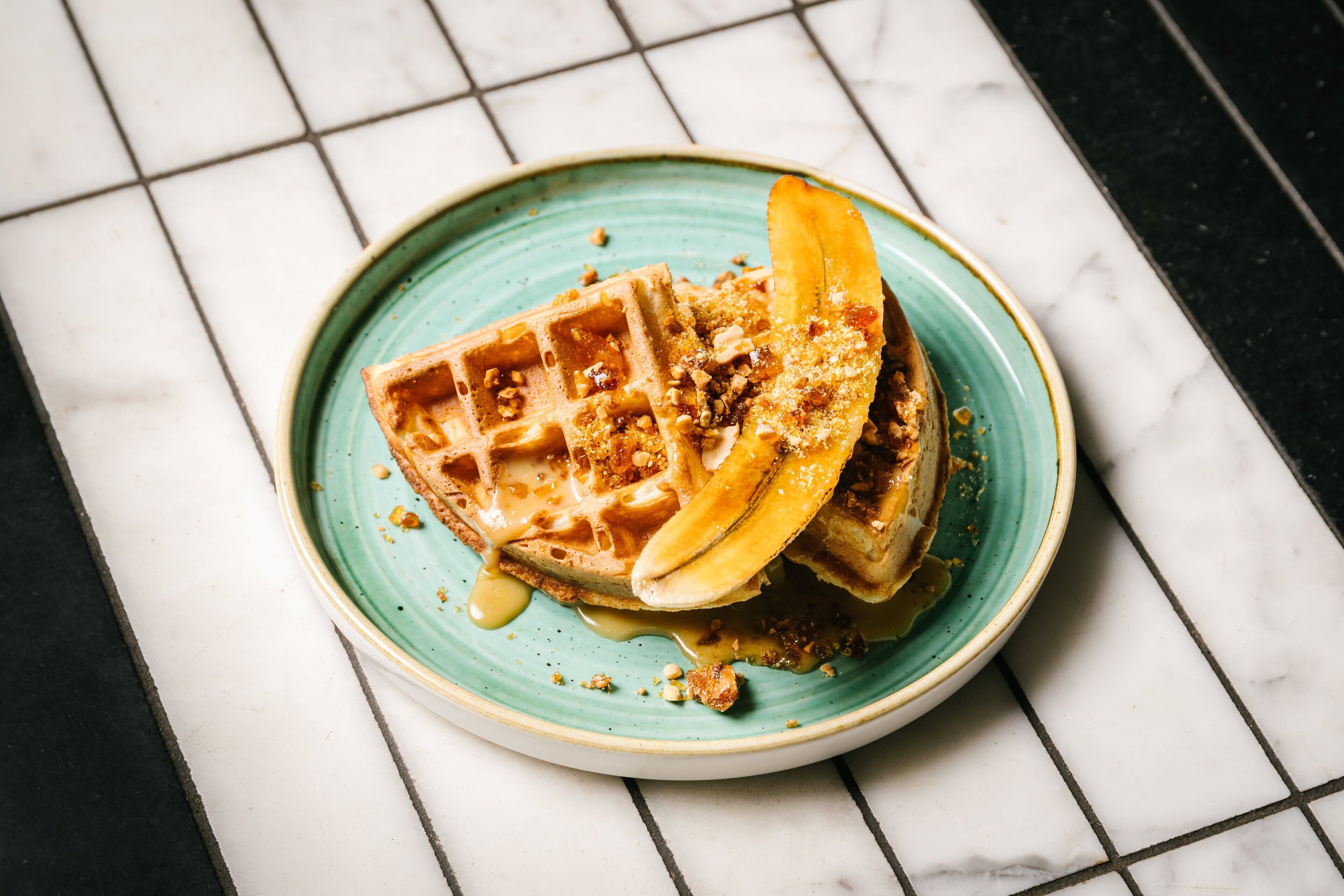 A plate with a waffle, banana slices, and syrup on a tiled surface.