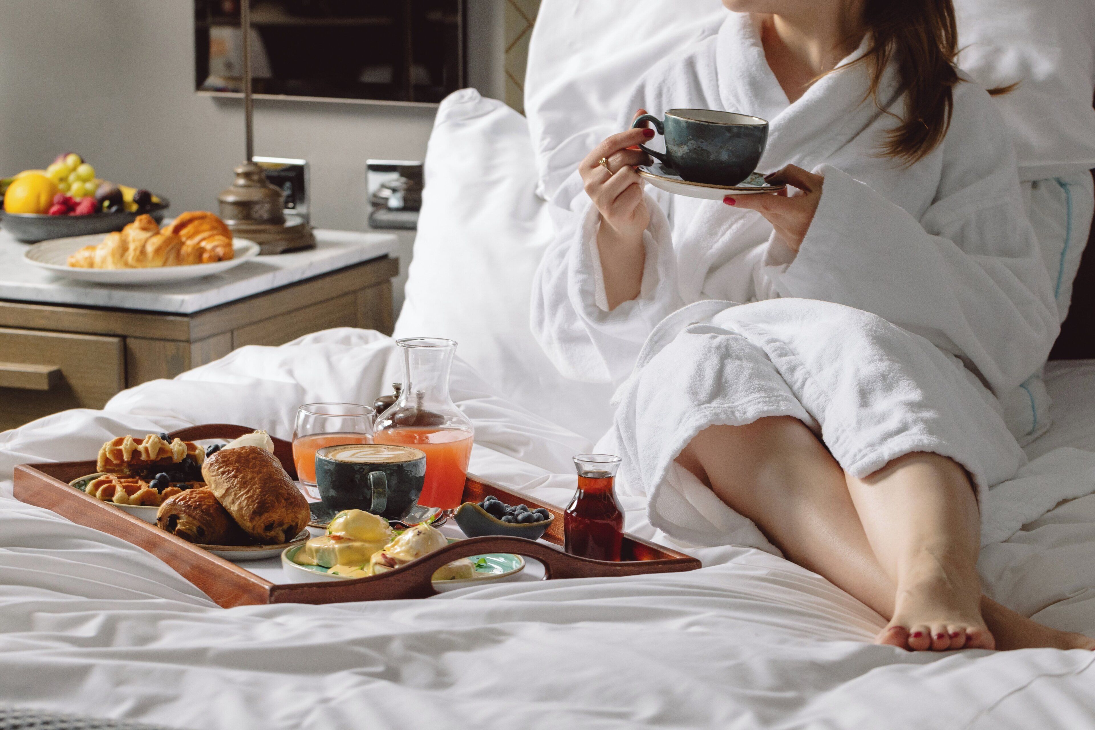 A person in a white bathrobe sitting on a bed with a tray of food, a glass of orange juice, and a cup of tea.