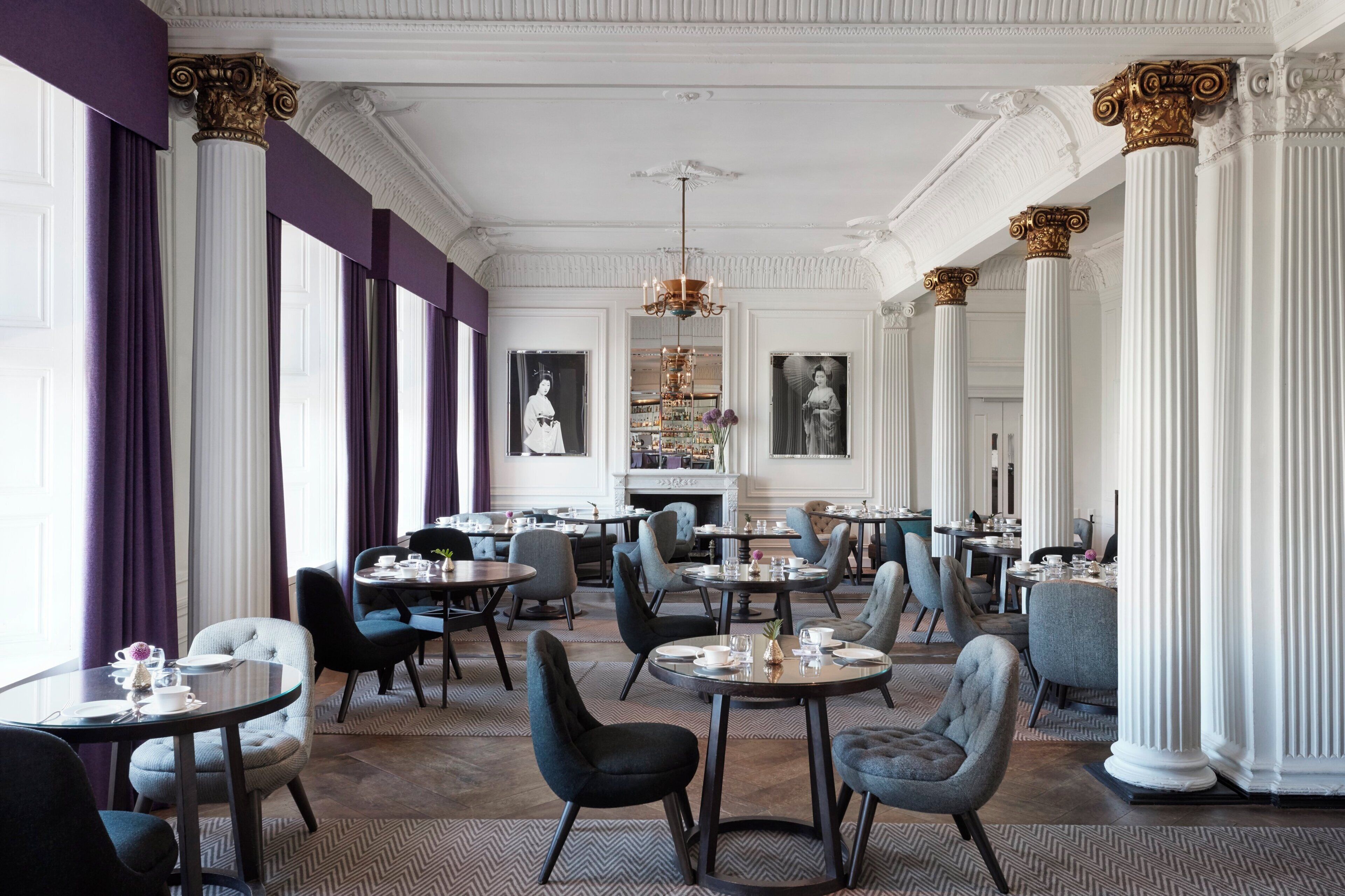 Un comedor espacioso con techos altos, columnas ornamentales y chimenea. Las mesas están dispuestas con vajilla y cristalería fina.