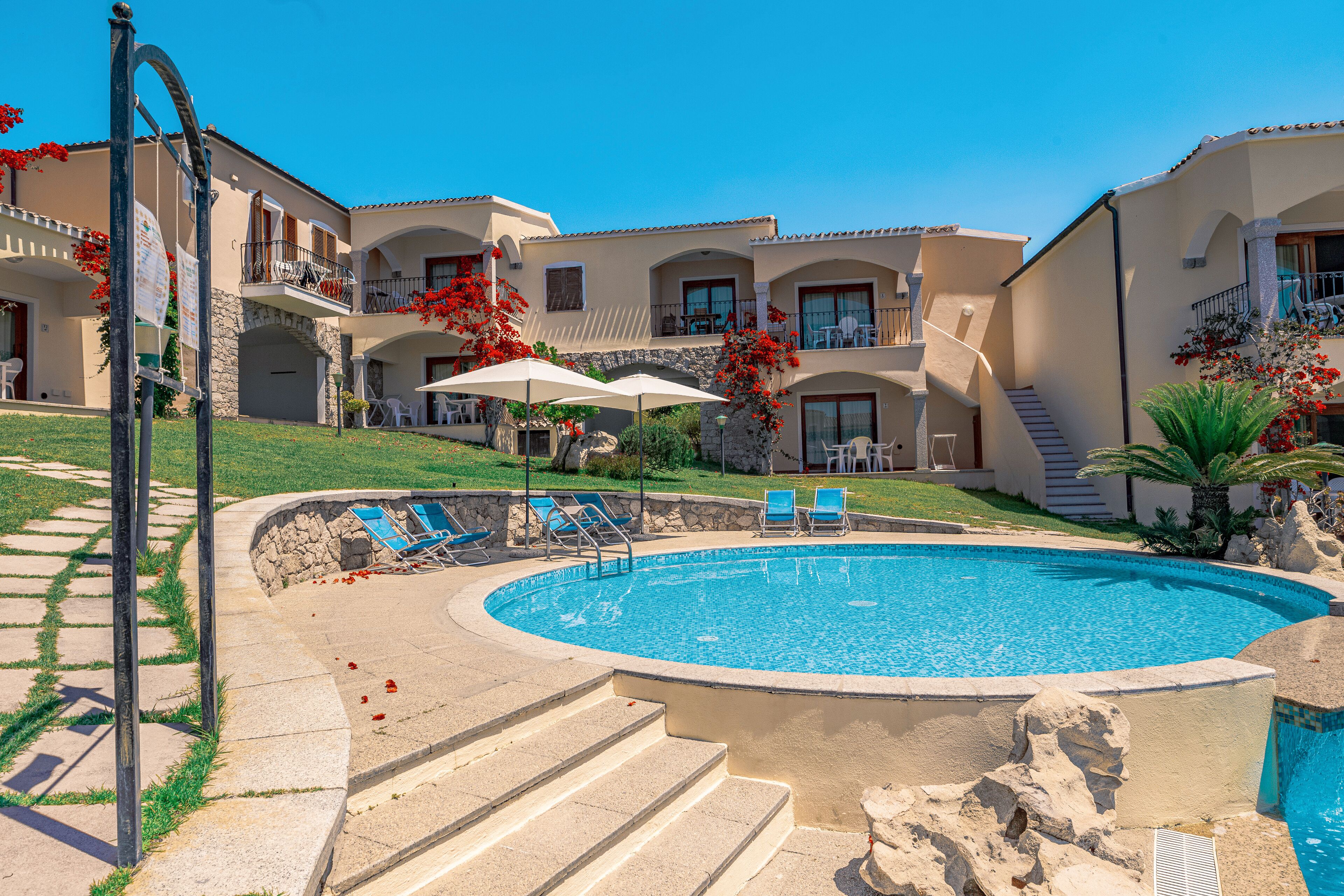 A swimming pool with lounge chairs and umbrellas, surrounded by a stone wall and steps leading to a building with multiple balconies.