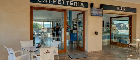 An outdoor seating area with a 'CAFFETTERIA' and 'BAR' sign, white chairs, and a tiled floor.