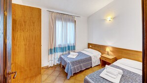 A hotel room with two beds, a wooden door, a window with curtains, a bedside lamp, and a wall-mounted light.