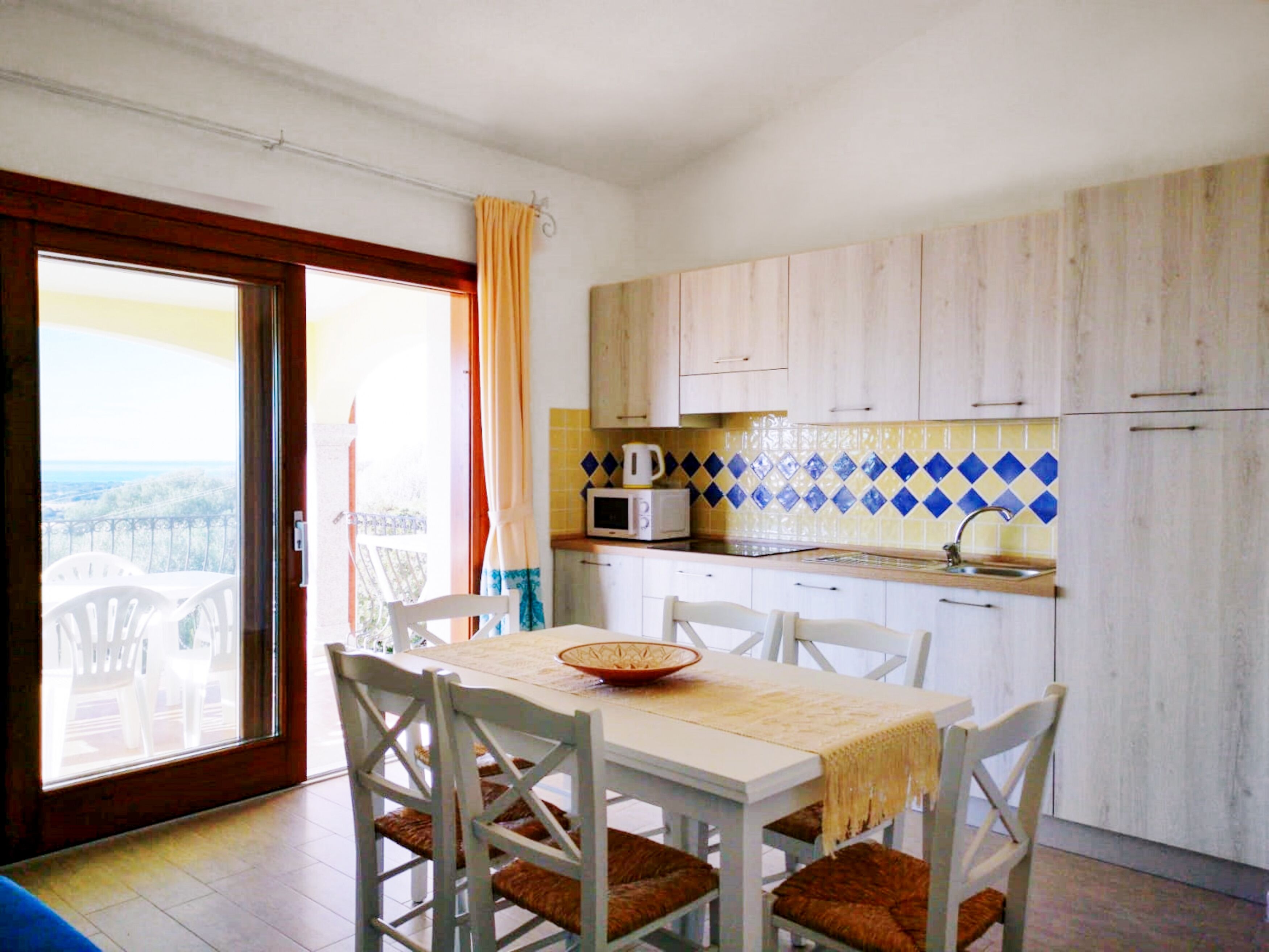 A dining area with a table and chairs, a kitchen with wooden cabinets and a microwave, and a view through a glass door.