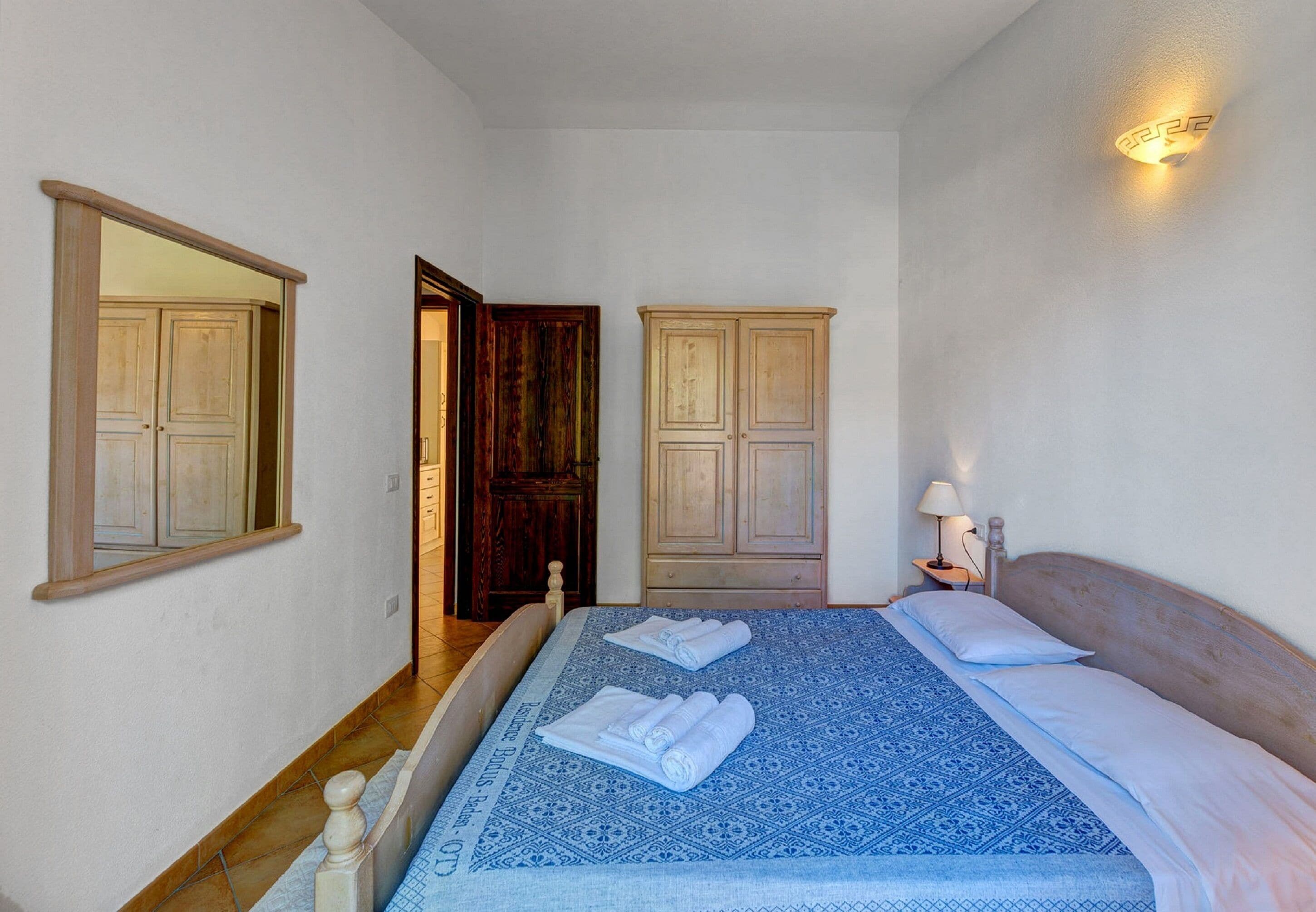A bedroom with a bed, two wooden wardrobes, a nightstand with a lamp, and a mirror on the wall.