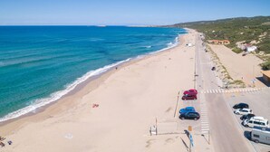 An aerial view of a beach with a road running parallel to the shore, lined with parked cars and a few people.