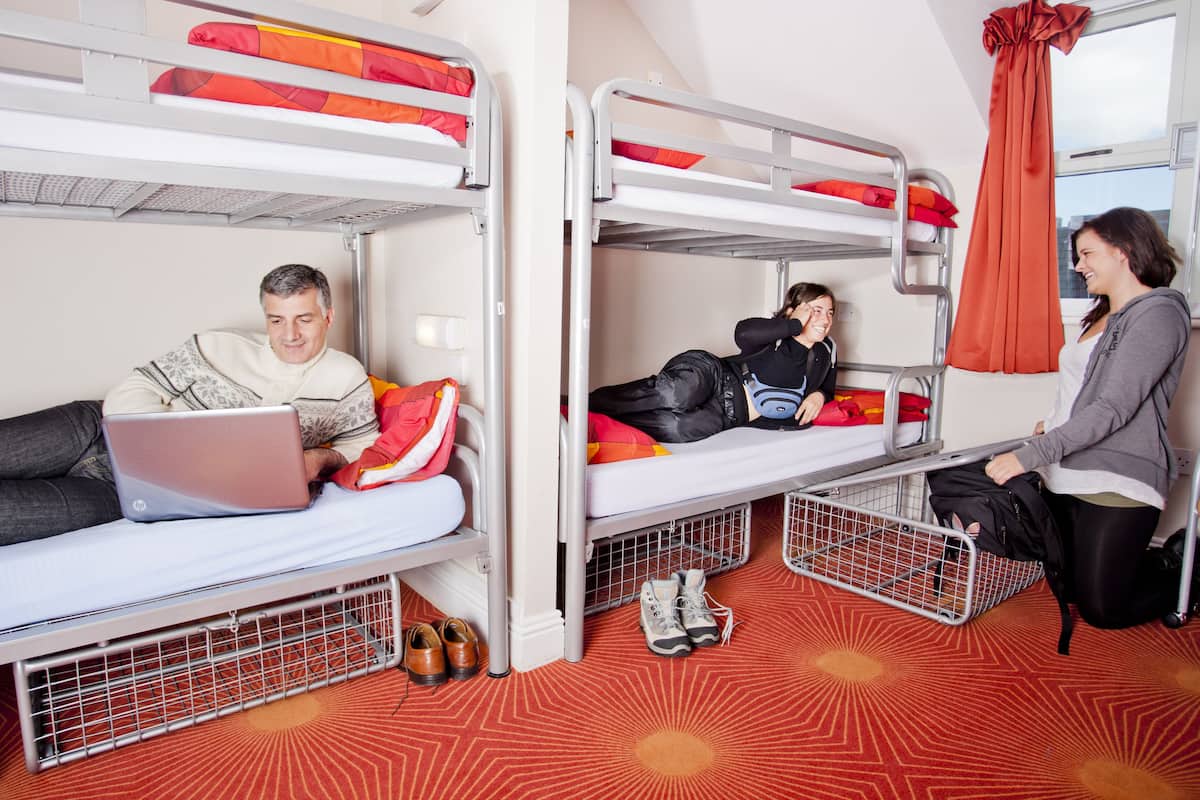 Ein Schlafsaalzimmer mit Etagenbetten, einem Laptop auf einem Bett und einer Person, die am Boden sitzt.